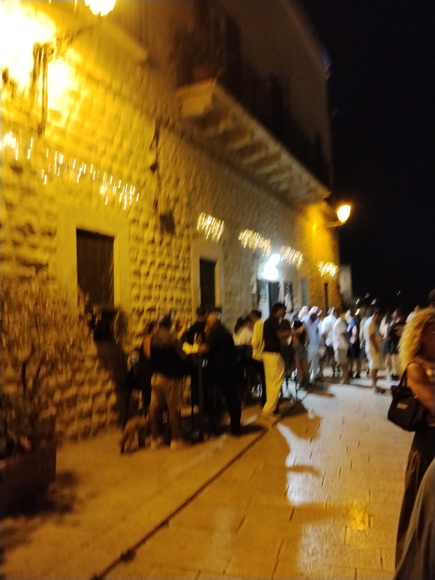 The image depicts a street scene at night in Bari, Italy, likely a pedestrian area with shops or restaurants. The architecture suggests an older European city, with a stone building on the left illuminated by strings of warm yellow lights and a lamp post. In the foreground, a planter with some vegetation is visible. A group of people are gathered, some standing and conversing, others potentially seated at tables that are out of focus. The ground appears to be paved with cobblestones or similar material. The overall atmosphere is one of a lively evening gathering.