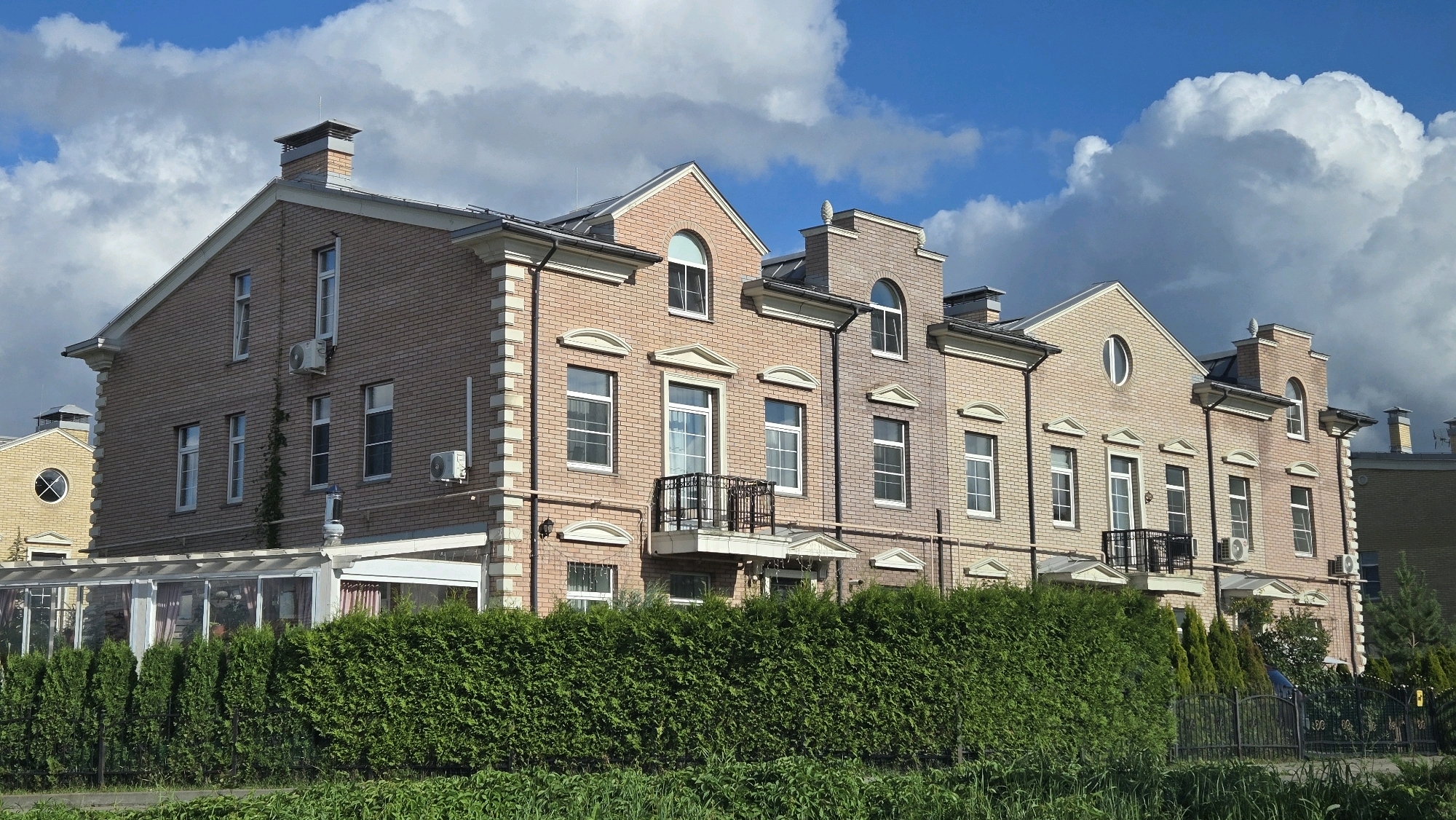 This is a frontal, eye-level, medium-distance, full-shot photograph of a row of attached houses in the city of Орлов Кп, Russia. The houses are constructed from light-colored bricks and feature pitched roofs. They have multiple windows, some of which are arched. Air conditioning units are visible mounted on the exterior walls. A dense, green hedge runs across the foreground, obscuring the lower portions of the houses. Behind the hedge, a black metal fence can be seen. The sky above is blue with large, white, puffy clouds. To the left, a greenhouse structure with a glass roof is attached to one of the houses. The overall impression is that of a well-maintained residential area. There are no people visible in the image.