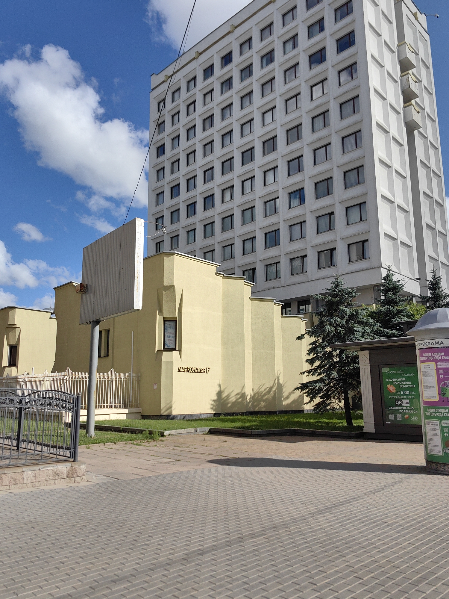 This image shows a view of a tall, white, rectangular office building with many windows. In front of it is a smaller, pale yellow building with a few windows. A large, metal advertising billboard stands in front of the yellow building. The buildings are located in Minsk, Belarus, specifically at 17 Masukova Street, as indicated by the Cyrillic text on the yellow building. The area in front of the buildings is paved with interlocking concrete bricks and there is a sidewalk. There is a metal fence in front of the yellow building. Several evergreen trees are visible between the yellow building and a kiosk or display stand on the right side of the image. The sky is blue with scattered white clouds. The scene is brightly lit by sunlight, casting shadows on the ground.