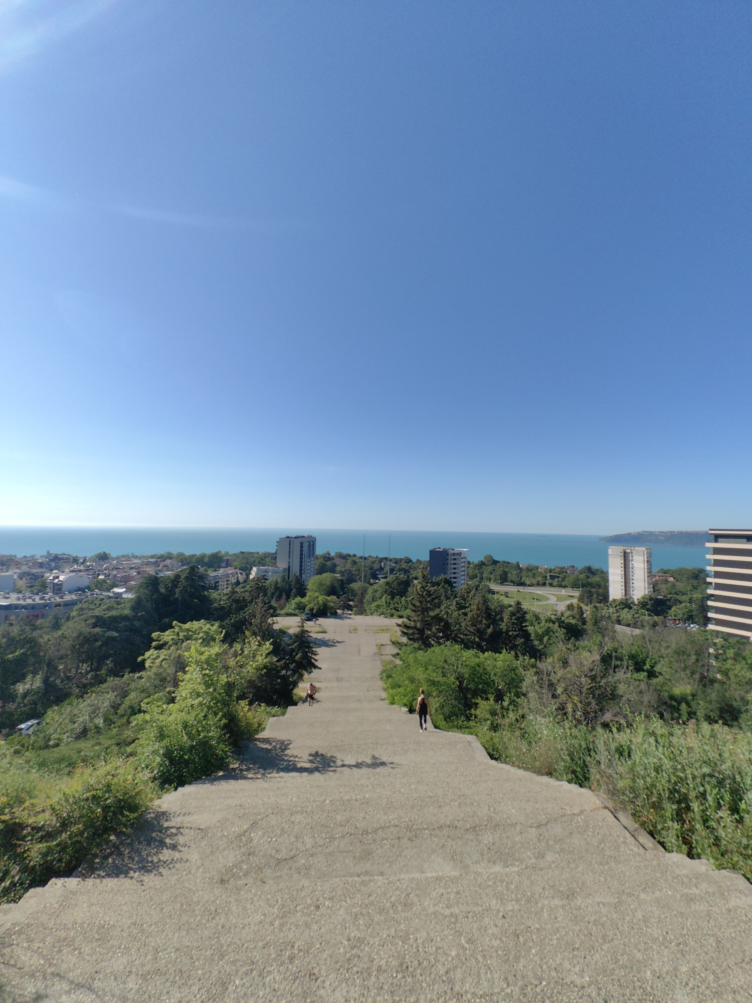 This is a wide-angle, eye-level shot looking down a wide, concrete staircase in Varna, Bulgaria. The staircase appears to be a part of a large outdoor structure, possibly an abandoned or disused monument or observation point, as it is unadorned and shows signs of wear. The staircase leads down towards a city visible in the middle ground, which is nestled between lush green trees. Beyond the city, a vast expanse of calm, turquoise water, likely the Black Sea, stretches to the horizon under a clear, bright blue sky. Several tall, modern buildings, typical of urban architecture, are interspersed among the lower structures of the city. Two individuals are visible on the staircase: one person is sitting further up and to the left, while another, a woman in athletic wear, is walking down the stairs away from the camera. The surrounding vegetation is dense and green, with trees and bushes growing on either side of the staircase and cascading down the slopes. The overall impression is one of a scenic urban overlook on a sunny day.
