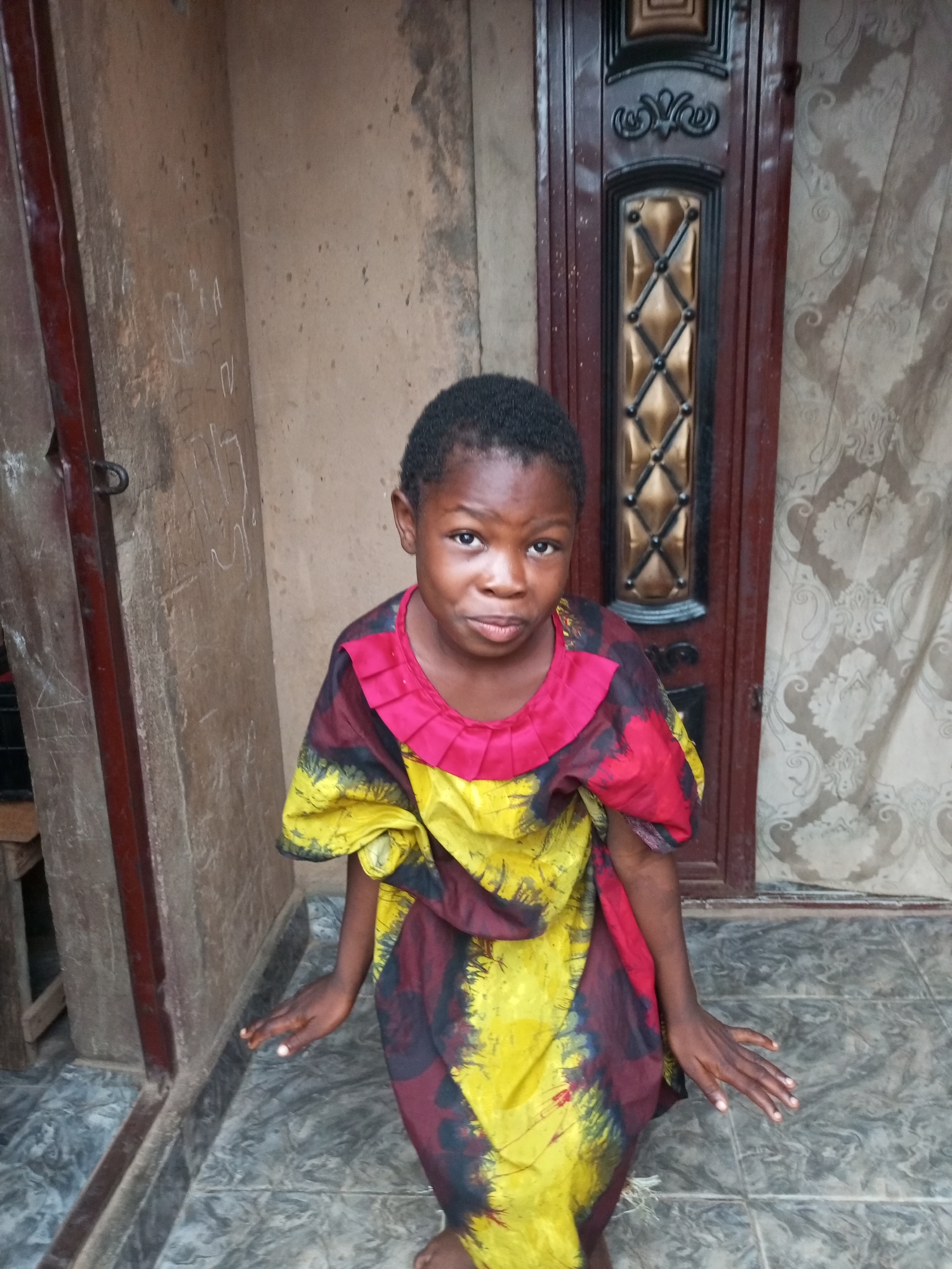 A young Black girl stands in front of a weathered, light brown wall and a dark brown door with ornate metallic detailing. She is wearing a colorful dress with yellow, red, and dark blue patterns. Her hands are slightly raised and open, palms facing outward. The floor is tiled with a grey and black marbled pattern. The overall environment appears to be an outdoor space, likely a courtyard or entryway, in a setting that suggests a developing urban area. The girl's expression is direct and curious. This scene is consistent with an outdoor setting in Tempure, Nigeria.
