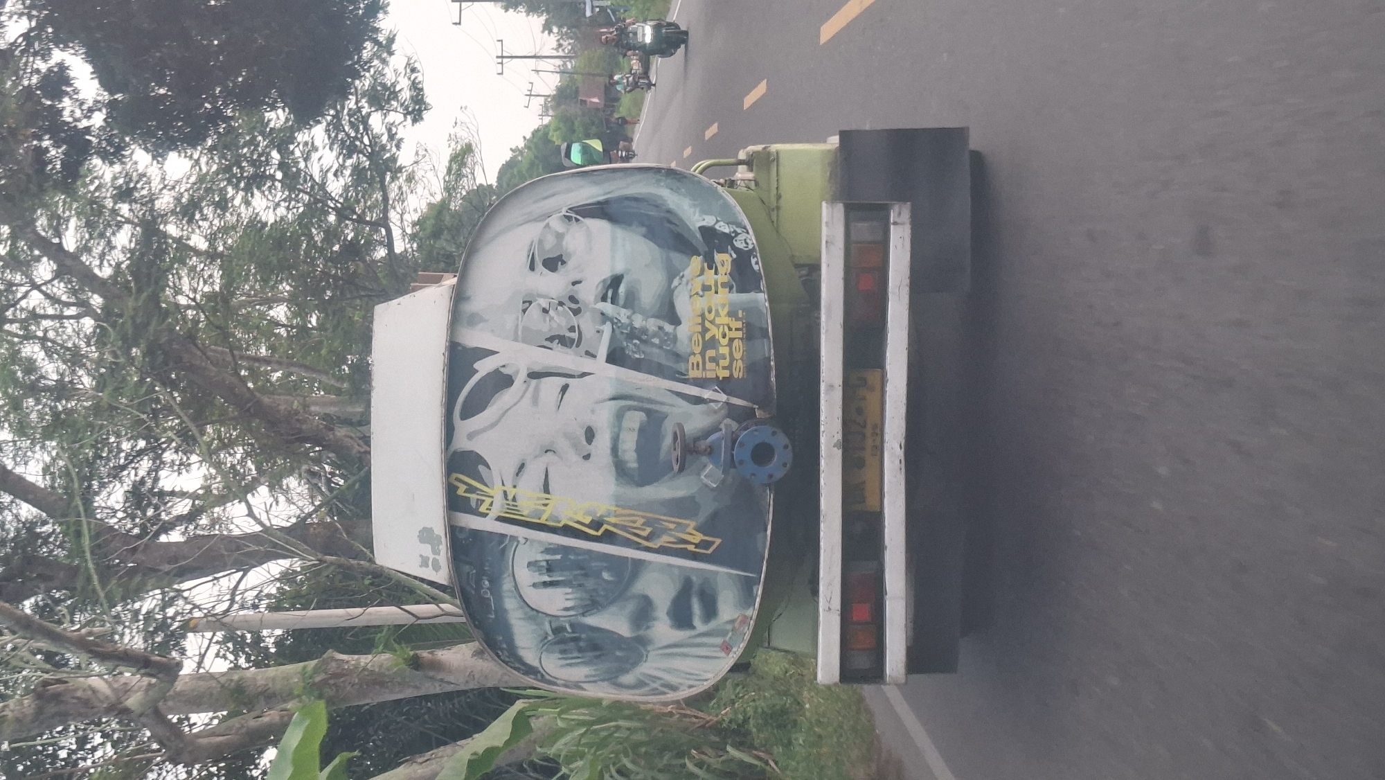 This image, captured on a road in Batu Malenggang, Indonesia, shows the rear view of a water tanker truck. The truck's cylindrical tank is adorned with a graphic featuring three stylized faces, two wearing large sunglasses and one with its mouth open in a wide laugh. The graphic has text that is partially obscured. A blue valve is mounted on the lower section of the tank. The truck has a yellow license plate with faded text. The road is paved with asphalt and marked with yellow lines. In the background, there are trees, utility poles, and a few motorcycles and their riders on the road. The sky appears overcast.