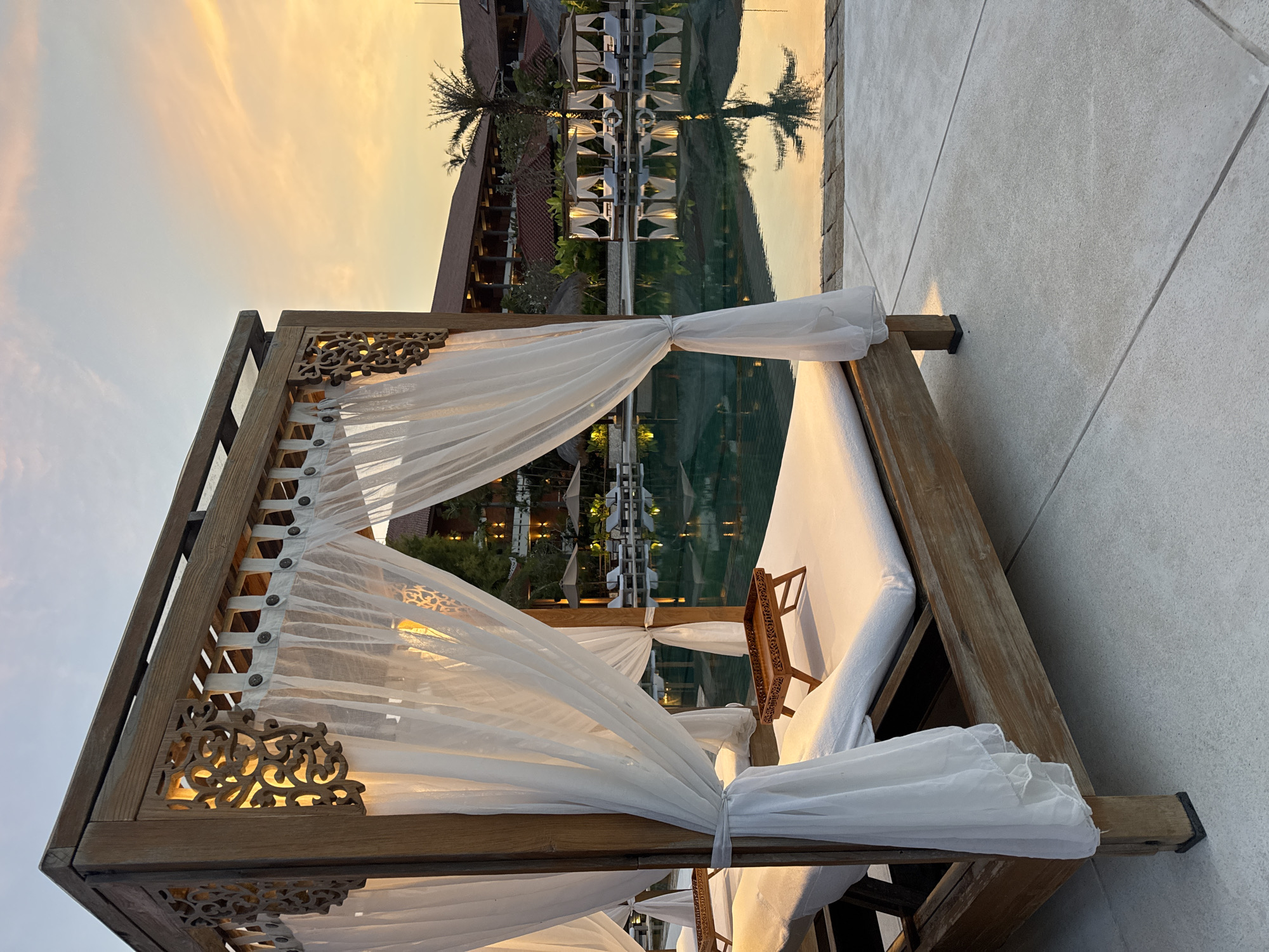 This image depicts a serene, unoccupied poolside area at a resort in Nusa Dua, Indonesia, during sunset. The foreground is dominated by a large, four-poster wooden cabana, featuring a rustic light-brown frame with ornate, intricately carved wooden panels on its upper corners, emitting a warm yellow light. White, sheer fabric curtains are tied back to the posts, revealing two white mattresses or daybeds within. A small, dark-brown, carved wooden tray with short legs rests on one of the mattresses. The cabana stands on a light grey concrete or tiled surface with visible expansion joints.
In the midground, a large swimming pool with calm, dark teal-green water reflects the warm sunset sky and the distant structures. Along the far edge of the pool, several identical wooden cabanas with white drapes are arranged, some accompanied by sun loungers and light-colored umbrellas.
The background features a multi-story resort building with traditional Indonesian architecture, including dark wooden elements and reddish-brown tiled roofs. Lush tropical foliage, including prominent palm trees, surrounds the buildings and pool area. The sky above transitions from a light blue at the zenith to vibrant orange and yellow hues near the horizon, indicative of sunset. No individuals are visible in the scene.