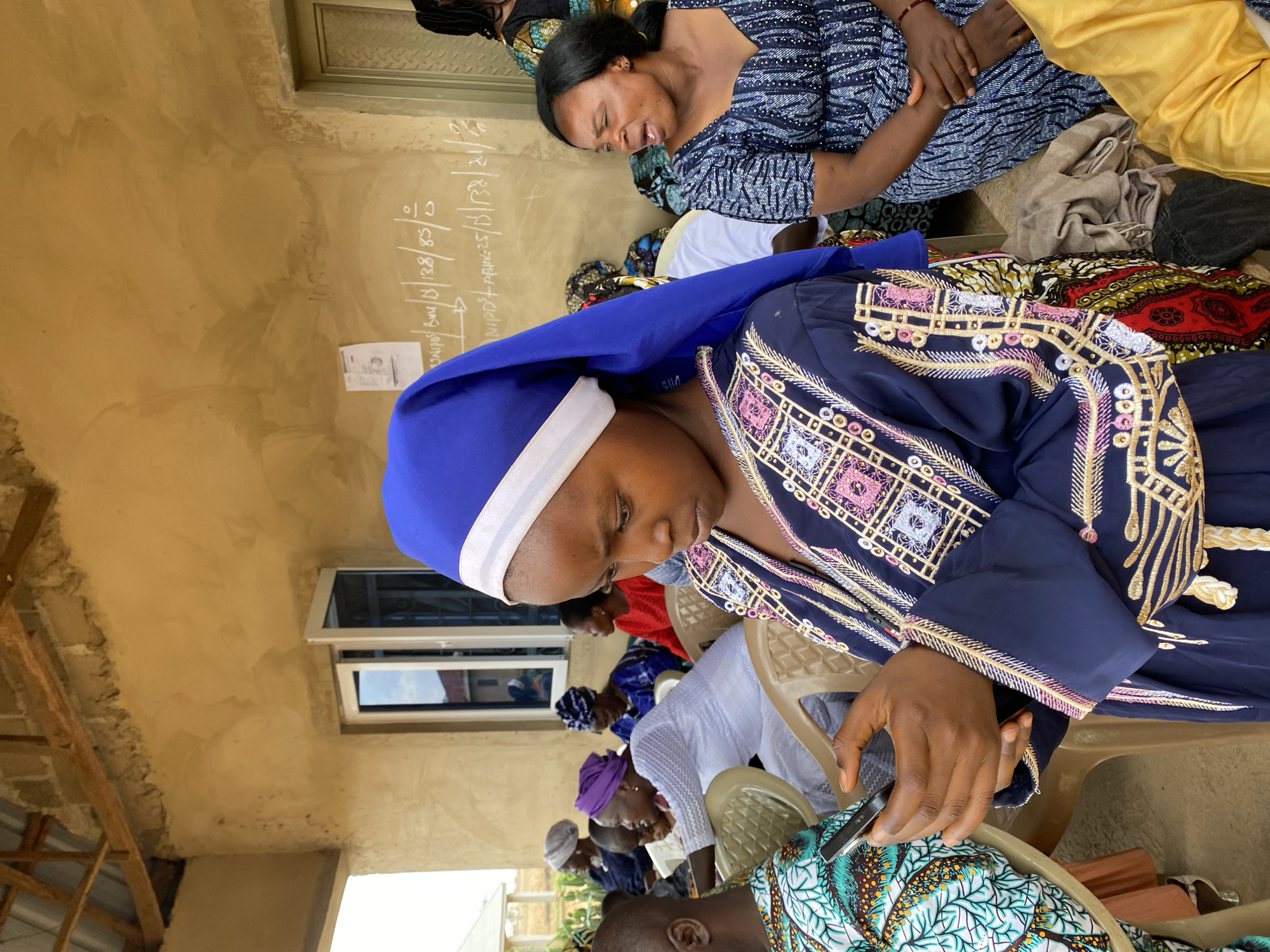 A medium shot depicts a gathering of individuals in what appears to be an interior space in Kuru Karama, Nigeria. The focal point is a woman wearing a navy blue robe adorned with intricate embroidery featuring geometric patterns in gold, pink, and white, along with a blue and white head covering. She is seated and focused on a mobile phone in her hand.
Additional people are present, seated in chairs, including a woman in a patterned blue dress and another in yellow attire. The environment includes a pale yellow wall with some writing and numbers on it. A window provides a glimpse of the outside. The ceiling appears to be unfinished or partially constructed, showing wooden beams and a concrete structure.