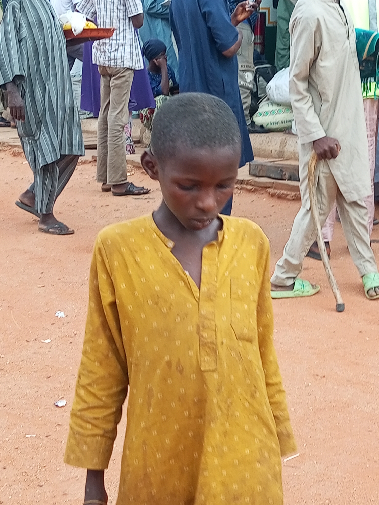 In Gombe, Nigeria, a young boy with dark skin and short, dark hair stands in the foreground, looking downwards. He wears a yellow, long-sleeved tunic with a repetitive, small, light-colored pattern. The tunic has a small pocket on the left chest and a few dark stains. Behind him, several people are partially visible, suggesting a public gathering or market scene. To the left, a person in a grey and white striped robe walks away, their legs and feet visible wearing dark sandals. Next to them, another person in a white striped shirt and light brown pants is also walking. Further to the right, a person in a light-colored tunic holds a walking stick, wearing light green sandals. Another person in a blue garment is visible in the background, and a dark blue fabric is draped over a person. The ground is covered in reddish-brown dirt.