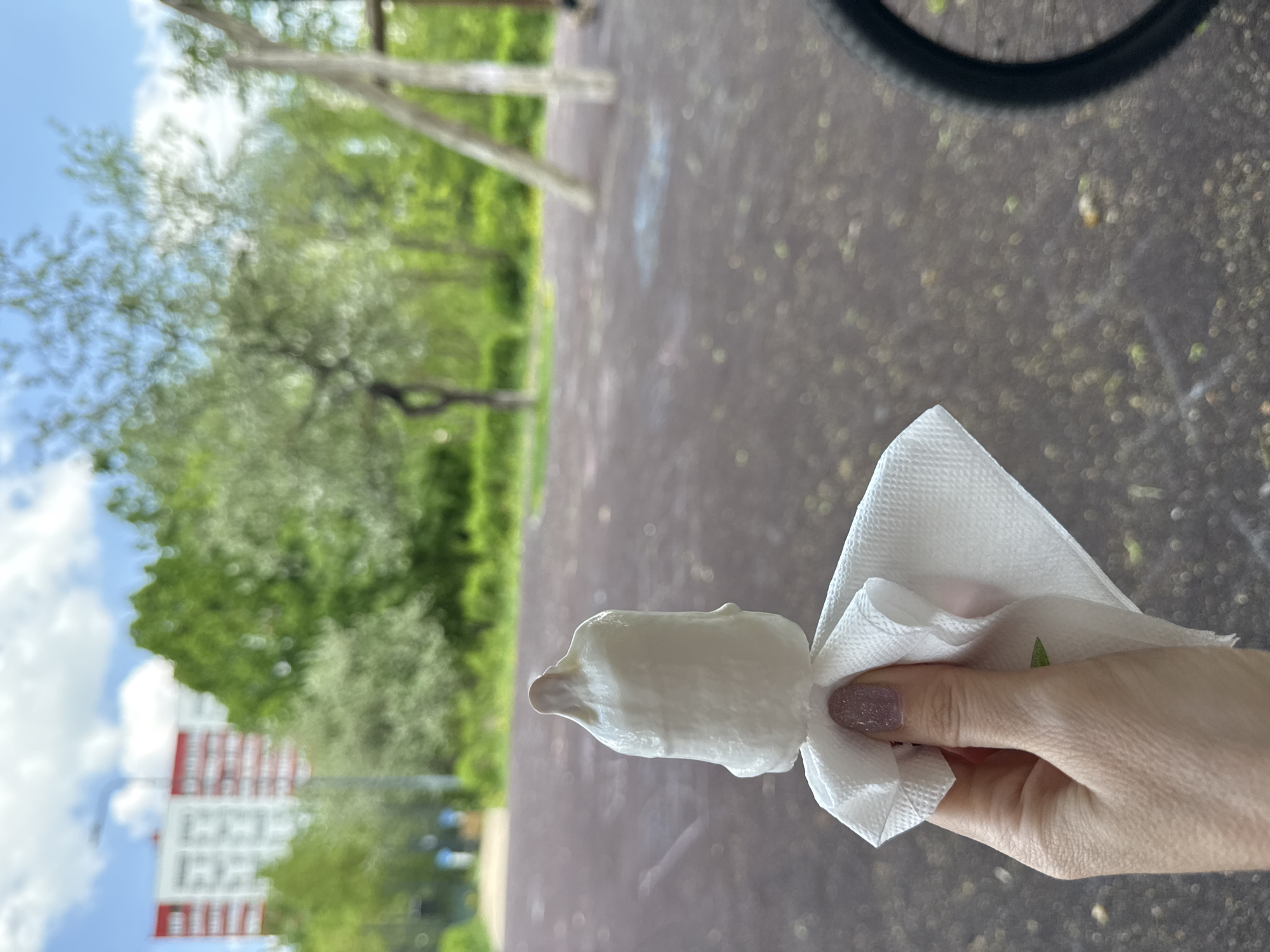 A medium shot depicts a partially eaten, white-colored ice cream treat held in a person's right hand, wrapped in a white paper napkin. The background suggests a park or outdoor recreational area. A blurry image of a bicycle wheel is visible in the upper right corner. Trees and greenery occupy the mid-ground. In the lower-left background, a multi-story building with white and red accents is partially visible. The image's perspective suggests it was taken in Moscow, Russia.