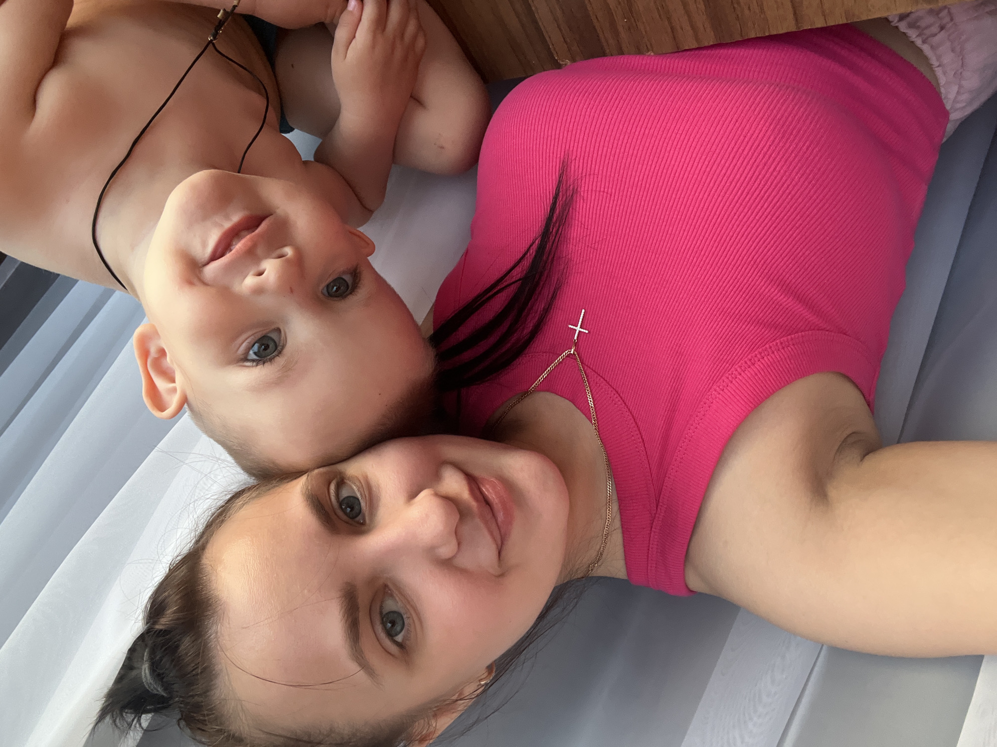 A woman and a baby are lying down next to each other. The woman is wearing a pink ribbed tank top and a gold necklace with a cross pendant. She has brown hair pulled back and is smiling. The baby is lying on its back and is looking at the camera. The baby has a thin, dark cord around its neck. The background is a light gray surface. The location is Марковский, Russia.