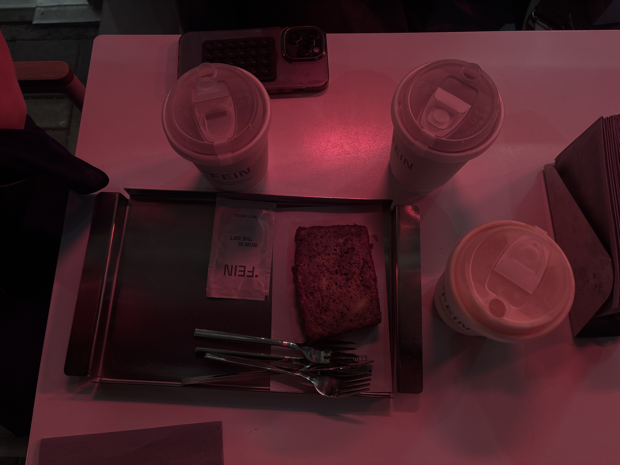 The image shows a table setting, likely in Yerevan, Armenia based on the "FEIN" branding visible on cups and a wet wipe. The setting consists of:
* Three disposable cups with lids, branded "FEIN."
* A silver metal tray containing a slice of dark bread, a sealed "FEIN" wet wipe packet, and three forks.
* A smartphone placed near the top left of the frame.
* A stack of napkins.
The lighting is dark and has a reddish tint. Part of a dark-sleeved arm is visible on the left edge, implying a person is seated at the table.