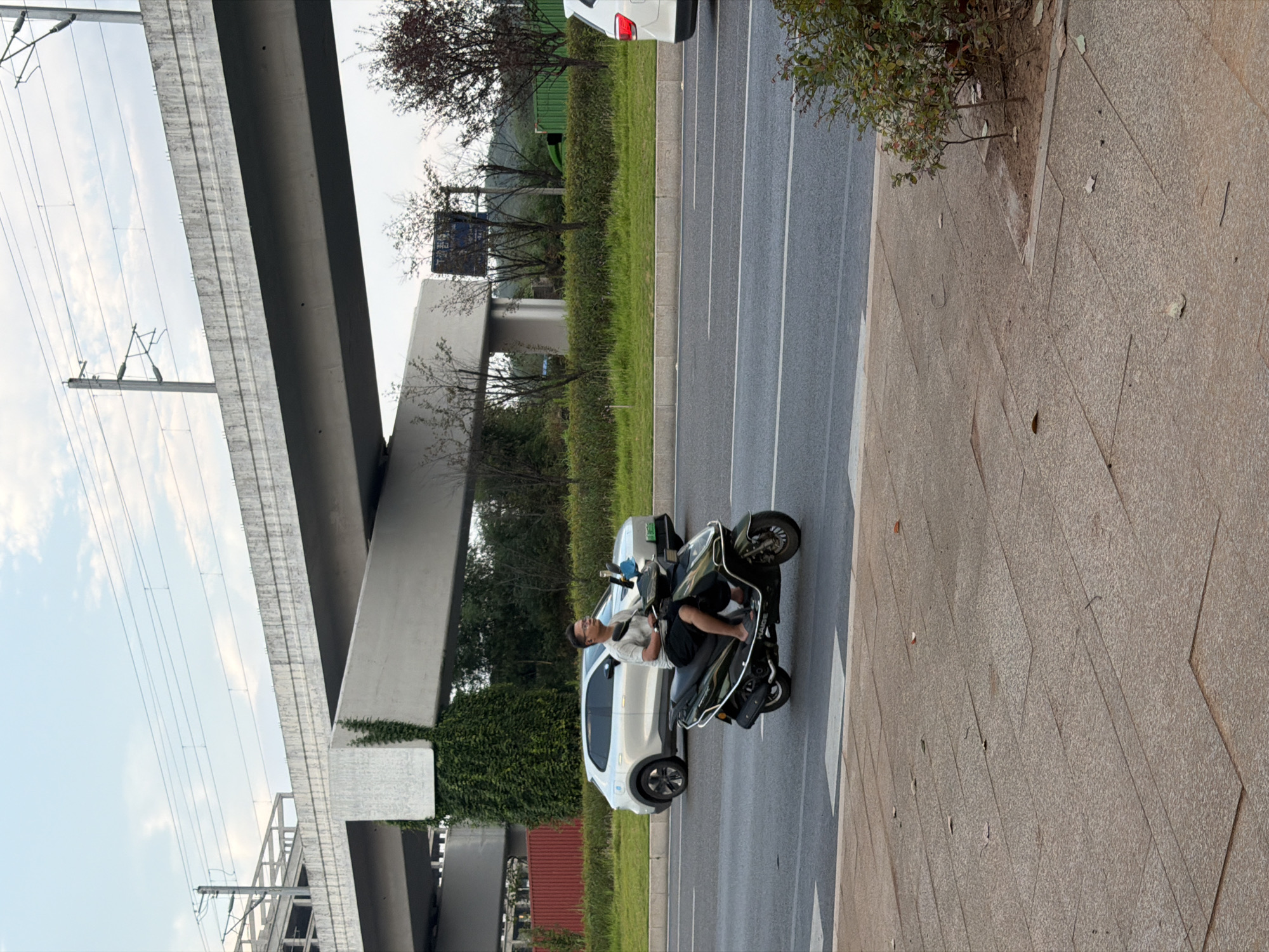 A man rides a black scooter on a paved road in Jiangjin, China. He is wearing glasses, a light-colored t-shirt, and dark shorts. The scooter has a silver trim. In the background, a silver SUV is parked on the side of the road. A large concrete overpass with electrical wires runs overhead. Lush green bushes and trees line the roadside, and a small tree is visible on the sidewalk in the foreground. The sky is cloudy.