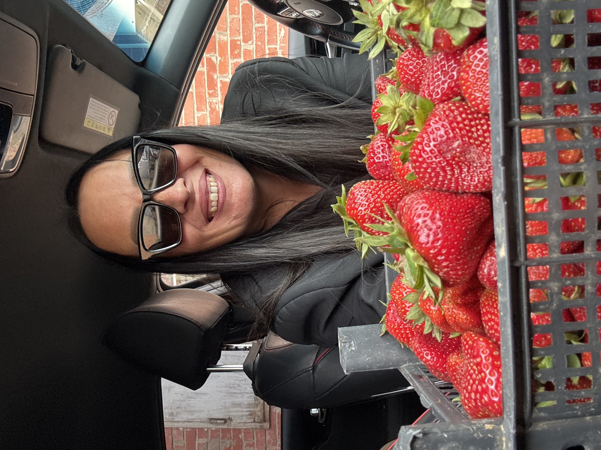 A woman with long dark hair and large black sunglasses is sitting in the driver's seat of a car. She is smiling and wearing a black blazer. In front of her is a large black crate overflowing with ripe strawberries. The car appears to be parked in an outdoor area with a brick building in the background. The location is the city of Chaikovsky, Russia.