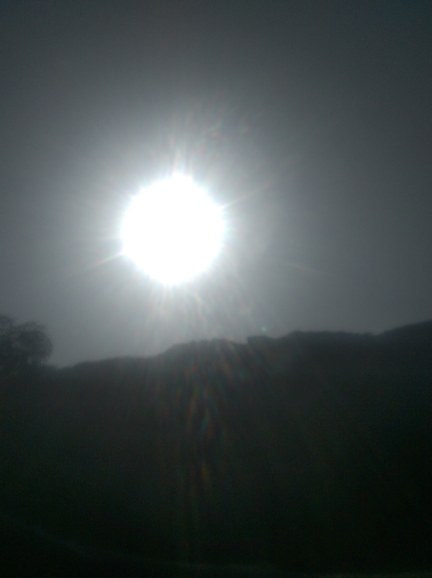 The image is a photograph of the sun at its zenith, appearing as a bright, spherical light source with radiating rays. The sun is positioned in the upper-middle section of the frame against a hazy, light-grey sky. Below the sun, a dark, silhouetted landscape is visible, suggesting hills or a treeline. The lower part of the image is almost entirely black, with some faint chromatic aberrations appearing as streaks of color, particularly on the right side. On the far left, a dark, indistinct shape suggests foliage or a tree. The overall impression is one of a bright day, possibly with some atmospheric haze, taken from an elevated or obscured viewpoint, characteristic of a hazy sky that can occur in Jalingo, Nigeria.