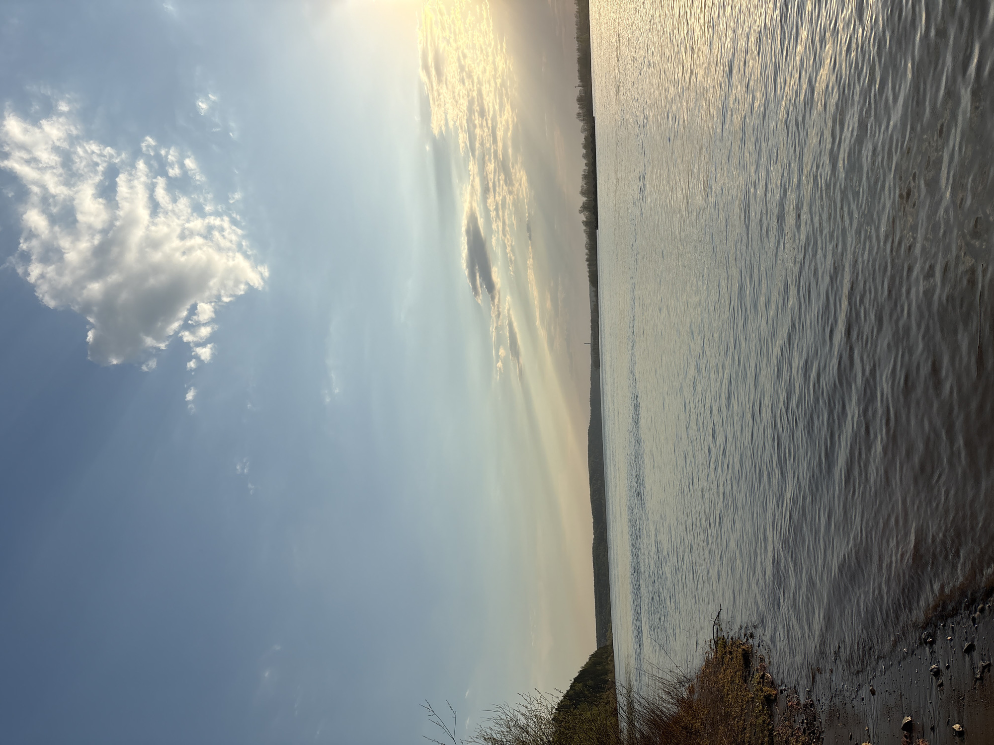 The image depicts a body of water, possibly a river or lake, under a partly cloudy sky. The sky is blue with scattered white clouds, with sunlight streaming through some of the cloud formations. The water surface reflects the sunlight, creating a shimmering effect. A shoreline is visible in the lower right corner, with vegetation growing along the edge. The horizon line appears to have a line of trees on the opposite side of the water. The image may have been taken at or near Ol'khovskoye, Russia, given the mention of the location.