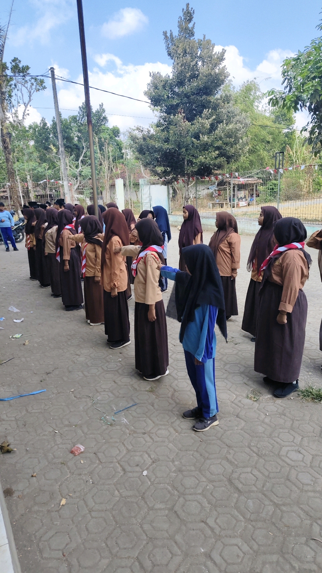 In the city of Banyubunih, Indonesia, a group of students in uniform are assembled outdoors on a paved area. The students, predominantly girls, are wearing uniforms consisting of brown skirts, light brown shirts, and hijabs in various shades of brown and black. Some students are wearing a red and white sash, indicating they are likely part of a scouting program.
The students are arranged in lines, facing towards the left of the frame. One student in the foreground, wearing a blue and black tracksuit over her uniform, is interacting with another student in the line by extending her arm towards her. The background shows trees, a fence, and some simple structures, suggesting a school or community gathering area. The sky is partly cloudy. The ground is paved with hexagonal concrete blocks, and there are some small debris and a blue string on the ground.