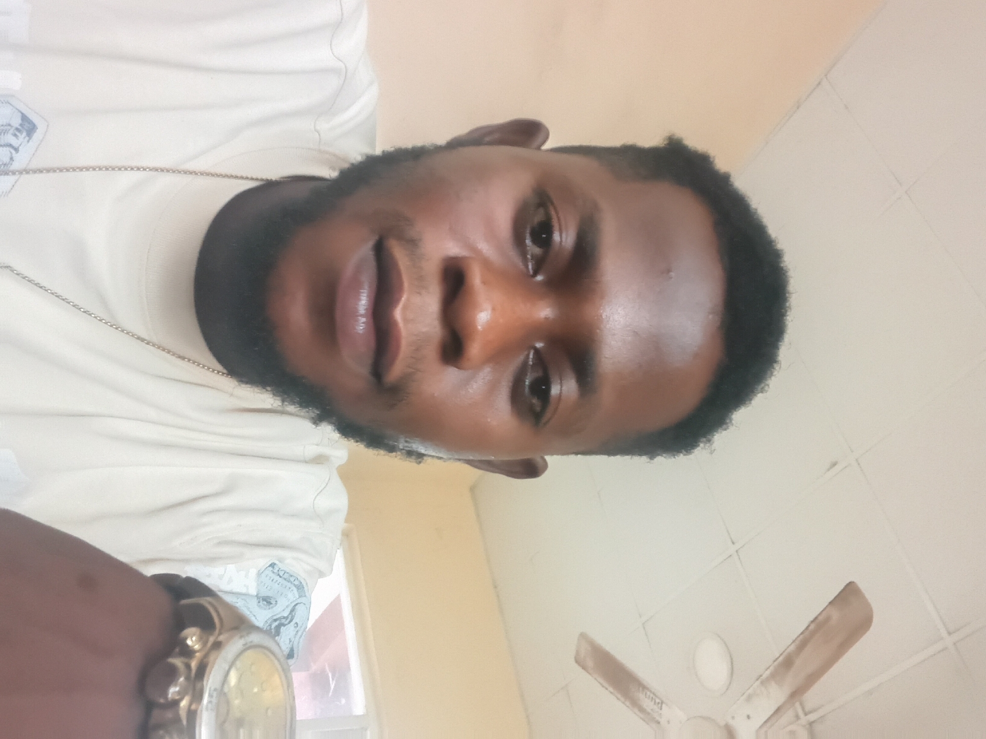 This is a selfie of a person indoors. The person is a man with dark skin and short black hair, and a beard. He is wearing a light-colored t-shirt and a gold chain necklace. He is also wearing a watch on his left wrist. The background appears to be a room with a light-colored wall and a tiled ceiling. There is a ceiling fan visible in the background. The lighting suggests it is daytime. The man's expression is neutral. There is no discernible activity or event taking place. No text is visible in the image.