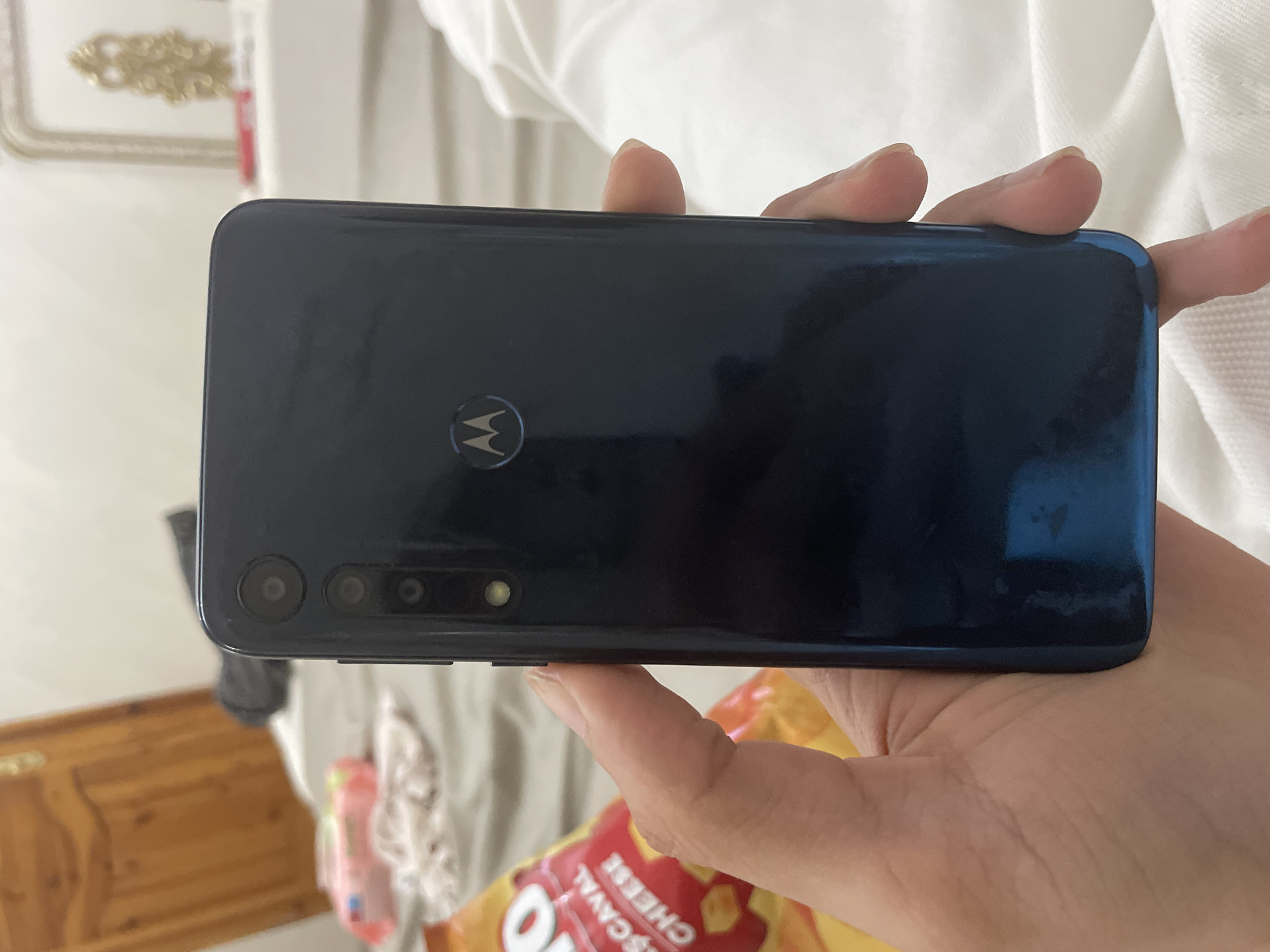 A person's hand holds a dark, glossy Motorola smartphone horizontally, showcasing its back panel. The Motorola "M" logo is centered on the phone's back. On the top left side of the device, a vertical camera module is visible, featuring at least three distinct lenses and an LED flash. The hand, with light skin tone, grips the phone, with fingers wrapped around the edges.
The background is an indoor setting, largely out of focus. In the upper left, a white surface with an ornate, golden decorative element, possibly part of furniture or wall decor, can be seen. The upper right background is dominated by white, textured fabric, consistent with bedding or a curtain. Towards the bottom left, a section of brown wooden furniture, possibly a dresser or cabinet, is visible. In the bottom right, a colorful snack bag, primarily red and yellow, is partially in view. The text "ȘCAVAL CHEESE" is clearly legible on the snack bag, indicating a cheese-flavored product, along with a partial letter "O". The lighting suggests it is daytime. The scene depicts a casual moment in an indoor environment.