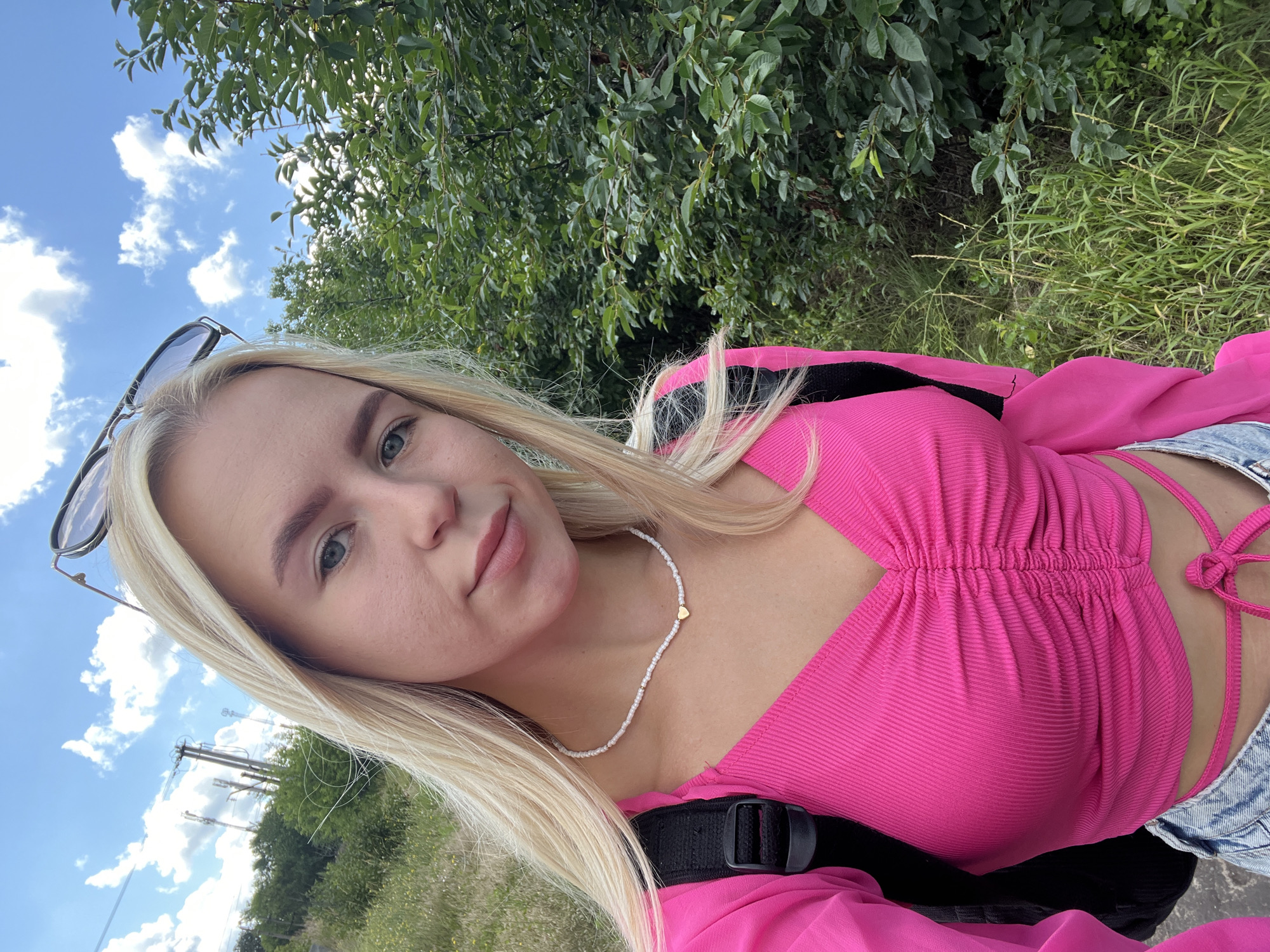A young Caucasian woman with blonde hair and blue eyes is taking a selfie outdoors. She is wearing a bright pink, ruched top with a V-neck and a white beaded necklace with a small gold heart pendant. She has a black backpack on. The background consists of green trees, tall grass, and a blue sky with scattered clouds. There are also power lines visible in the distance. The image appears to be taken during the daytime.