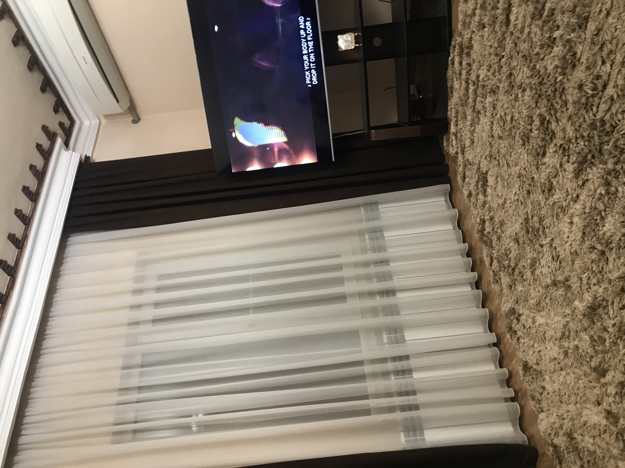 The image shows an interior view of a room. A window is covered by white horizontal blinds. A dark-colored drapery is positioned above the blinds. A television is mounted on the wall. A shelving unit is visible beside the television. A beige carpet is located on the floor. An air conditioning unit is mounted near the ceiling. Parts of a decorative wall molding are visible. It is not possible to determine the specific location in Yerevan, Armenia from the image alone.