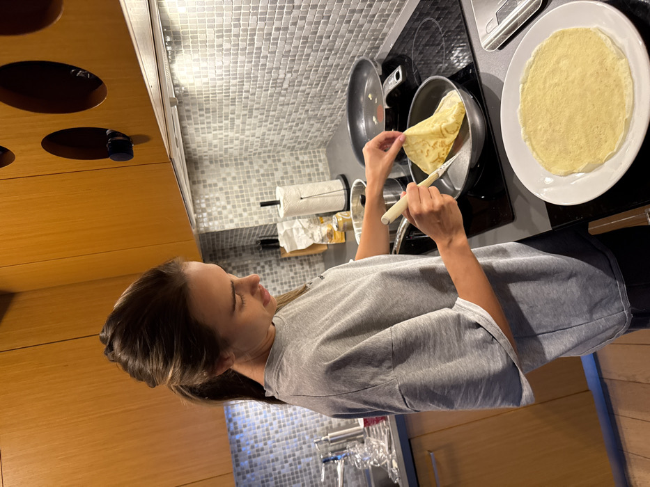 The image depicts a woman cooking in a modern kitchen. The subject, a female with dark hair tied back in a ponytail, is positioned in profile, facing right and looking down at a stovetop. She wears a grey, oversized short-sleeved t-shirt and dark trousers. Her left hand is holding the edge of a light-colored crêpe or thin pancake as it cooks in a dark frying pan on a black electric induction stovetop. Her right hand holds a light-colored spatula beneath the crêpe. A second dark frying pan is visible on a rear burner of the stovetop. To the right, on the dark countertop, a white round plate holds a single cooked crêpe, and a silver and black digital kitchen scale is partially visible.
The kitchen features light wood-grain upper and lower cabinets. The upper cabinets incorporate circular cutouts. Below the upper cabinets, a light grey mosaic tile backsplash covers the wall, illuminated by under-cabinet lighting. Behind the stovetop, a white paper towel roll on a dark stand is present, alongside other kitchen items including a small metal pot and jars. To the far left, a stainless steel sink area with a faucet is visible, with clear items drying in a rack. The flooring appears to be light-colored wood.
The image does not contain any discernible features, landmarks, or contextual information that would allow for the identification of a specific location within the city of Warsaw, Poland.