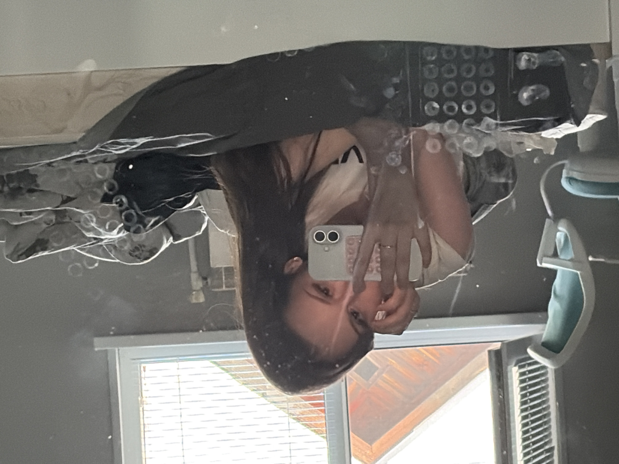 The image depicts an indoor scene specified as being in Semyonov, Russia, although no visual information within the image itself directly confirms this geographical location. A young female subject with long, dark brown hair is lying prone on a bed, capturing a mirror selfie with a light-colored smartphone held in her left hand. Her left hand obscures the lower half of her face, while her visible right eye looks towards the phone. She wears a light-colored t-shirt, and a thin ring is visible on her left ring finger.
The background, a reflection in the mirror, shows a light gray wall and a large window with white framing. The left side of the window has a partially open white louvered blind, while the right side features closed horizontal blinds, allowing strong natural light into the room. Outside the window, the reflection reveals a sloped roof with reddish-brown shingles, indicative of another building. A white radiator or heating element is faintly visible on the wall below the window.
To the far left, a modern office chair with a white frame and light blue-teal upholstery is positioned. In the lower foreground, the bed is covered by a dark gray fitted sheet and a light-colored patterned duvet, with some dark clothing draped on top. On a surface in the lower left, a dark rectangular storage box with multiple circular compartments is visible, topped with crumpled tissues. Numerous small, circular smudges and specks are present across the mirror's surface, particularly prominent in the lower half of the frame. The scene is brightly illuminated by natural light.
