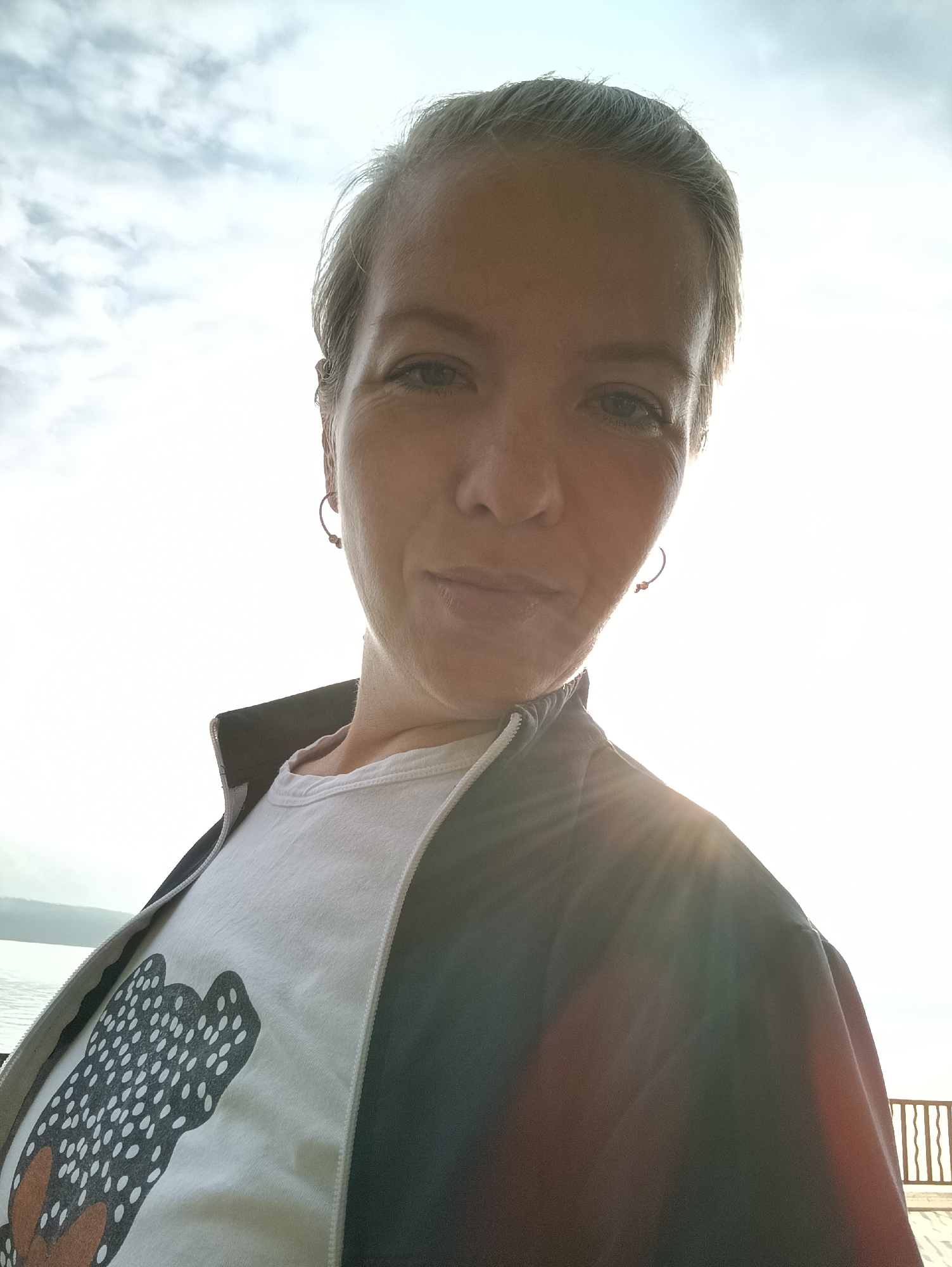 A close-up, low-angle selfie of a woman with short, graying hair and a light complexion. She is wearing a white t-shirt with a black and white polka-dot graphic of a butterfly, and a dark jacket. The woman is looking directly at the camera with a slight smile. The background is a bright, overcast sky with some clouds visible. A faint sun flare is present on the right side of the image. The location is on a waterfront promenade in Chaikovsky, Russia, with a railing visible in the lower right corner and a body of water on the left.