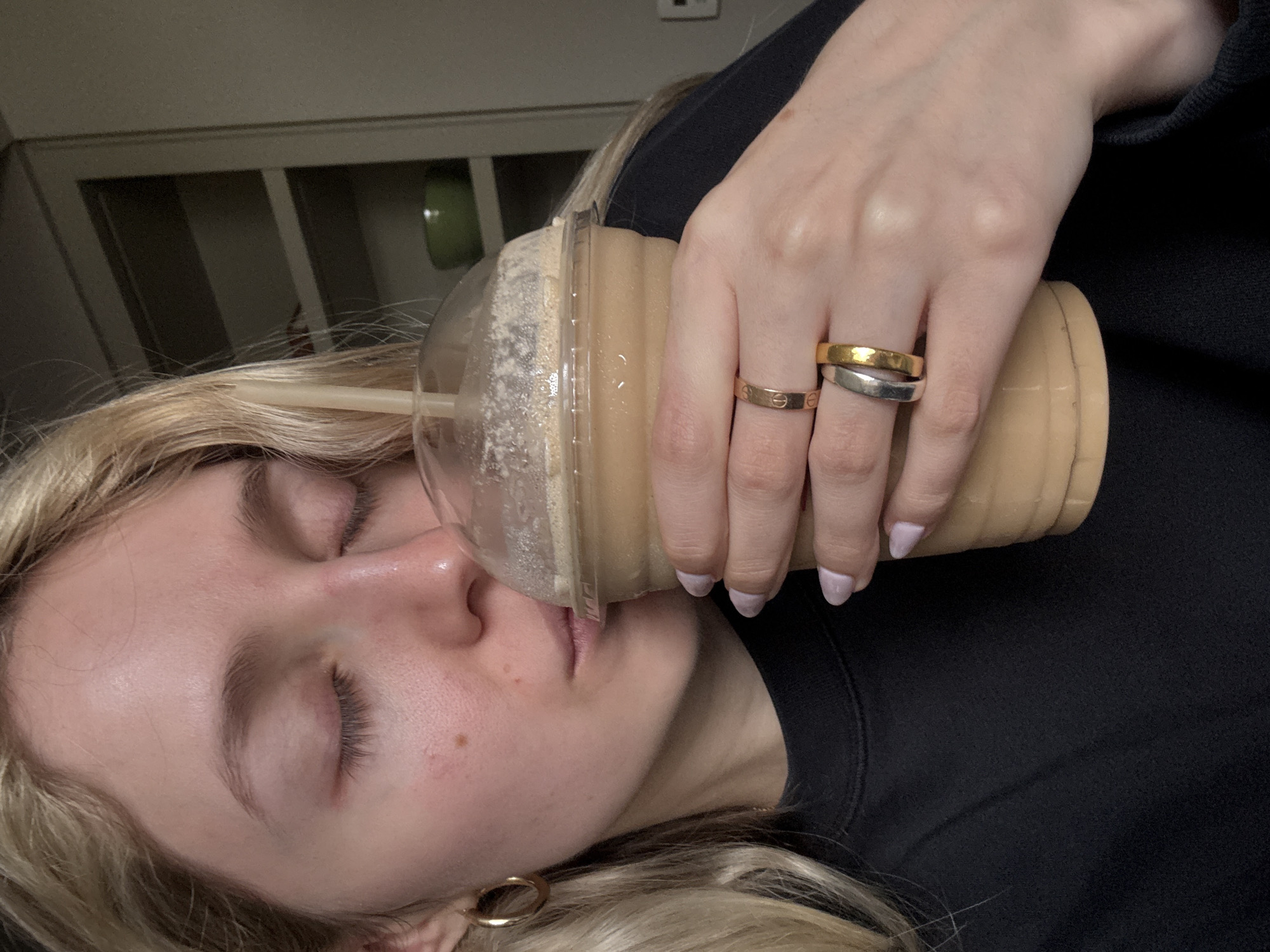A woman with blonde hair is resting her cheek against a large plastic cup filled with a light brown beverage, presumably a coffee drink. The cup has a clear dome lid and a straw. She is wearing a black sweater and gold hoop earrings. Her fingers are adorned with multiple rings, including a gold band, a two-tone (silver and gold) band, and a ring with repeating screw motifs. The background appears to be a room interior with a gray cabinet or shelving unit. The location in Mexico City, Mexico, cannot be verified from this image alone.