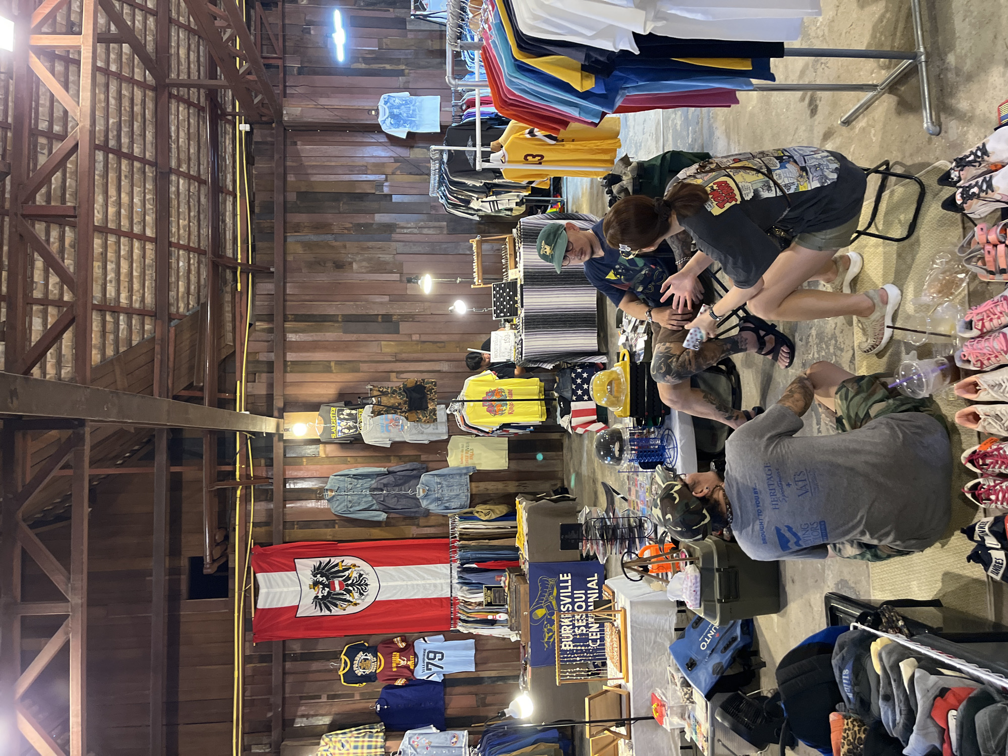 The image depicts an indoor market scene, likely a vintage clothing stall, within a structure with exposed wooden beams and paneling. The space appears to be a warehouse or a similarly rustic building, with a high ceiling.
In the foreground and midground, several people are engaged in conversation or browsing. A man wearing a camouflage baseball cap and a grey t-shirt with text on the back is seated on the floor. Beside him, a woman with her hair in a ponytail, wearing a t-shirt with a graphic print, is seated on a small folding chair. A man wearing a green baseball cap and a t-shirt with a graphic is also seated, facing the woman.
The stalls display a variety of clothing items, including t-shirts, jackets, and pants, hung on racks or folded on tables. There are flags, including an Austrian flag with an eagle emblem, displayed prominently. Various accessories and merchandise, such as jewelry displays, hats, and shoes, are also visible.
In the background, more racks of clothing are organized, and some mannequins are present. The overall atmosphere suggests a casual and bustling marketplace. The presence of an Austrian flag, along with numerous clothing items and what appear to be market stalls, indicates a commercial setting possibly catering to a diverse clientele or featuring international retro fashion. The lighting suggests it might be late afternoon or early evening, with some artificial lighting supplementing natural light entering from possibly unseen windows. The specific location within Chiang Mai City Municipality, Thailand, cannot be definitively determined from the visual cues alone, but it is consistent with the types of indoor markets and vintage clothing stalls found in areas like Nimmanhaemin or other artisan districts of the city.