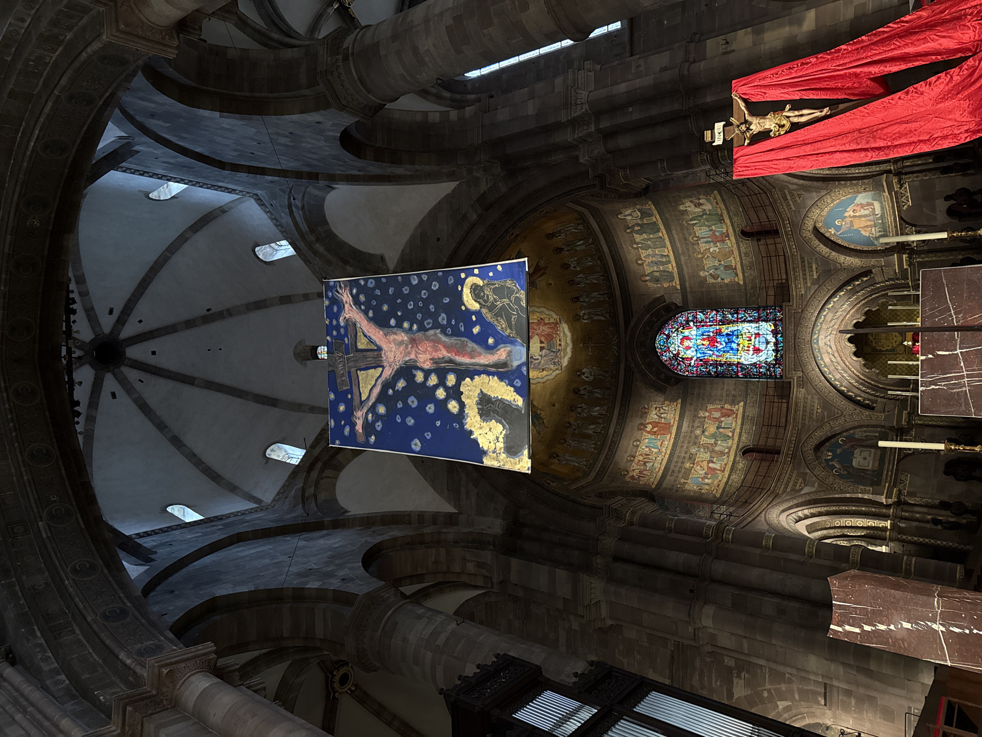 The image depicts the interior of the Strasbourg Cathedral, located in Strasbourg, France, specifically capturing the view towards the apse and choir from a low-angle perspective. The scene is dominated by massive, dark stone pillars and intricate ribbed vaults that form the cathedral's high ceilings. Several small, arched windows in the upper dome-like vault allow natural light into the otherwise dimly lit space.
A large, rectangular, contemporary painting is suspended centrally from the ceiling. This artwork features a stylized, reddish-skinned crucified figure with outstretched arms against a dark blue background punctuated by numerous small, luminous yellow and white spots. In the lower left of the painting, a dark-robed figure with a golden halo looks upwards, while a dark, horse-like shape is visible in the lower right.
Below this suspended artwork, the apse wall displays a vibrant stained-glass window, predominantly blue, red, and yellow, illustrating a central religious figure. The curved wall surrounding the window is adorned with multiple tiers of mosaic or fresco artwork, depicting rows of figures against a golden backdrop. To the right of the stained glass, a traditional gilded crucifix, featuring a figure of Jesus, is heavily draped with extensive swathes of bright red fabric that cascade to the floor. In the foreground, sections of dark, veined marble structures are visible, along with a single, tall white candle set in a golden holder. The environment presents a blend of ancient architecture and modern artistic intervention.