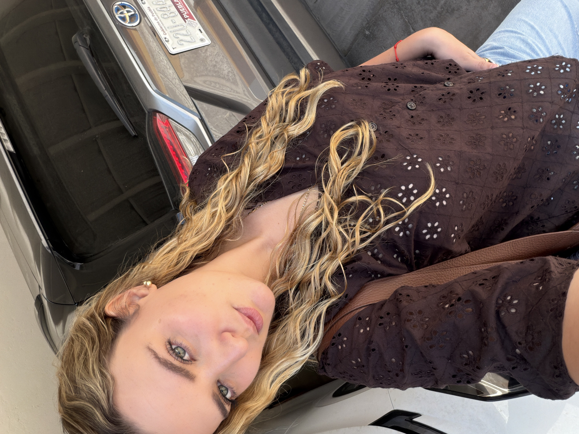 A waist-up shot depicts a fair-skinned woman with light brown hair, wearing a brown, floral-patterned shirt and blue jeans. A brown leather purse strap is visible across her shoulder. She is positioned in front of a car, with the license plate indicating it's from the state of Puebla, Mexico. The license plate number is 220U-844. The specific location within the city of San Bernardino Tlaxcalancingo, Mexico, is not discernible from the image.