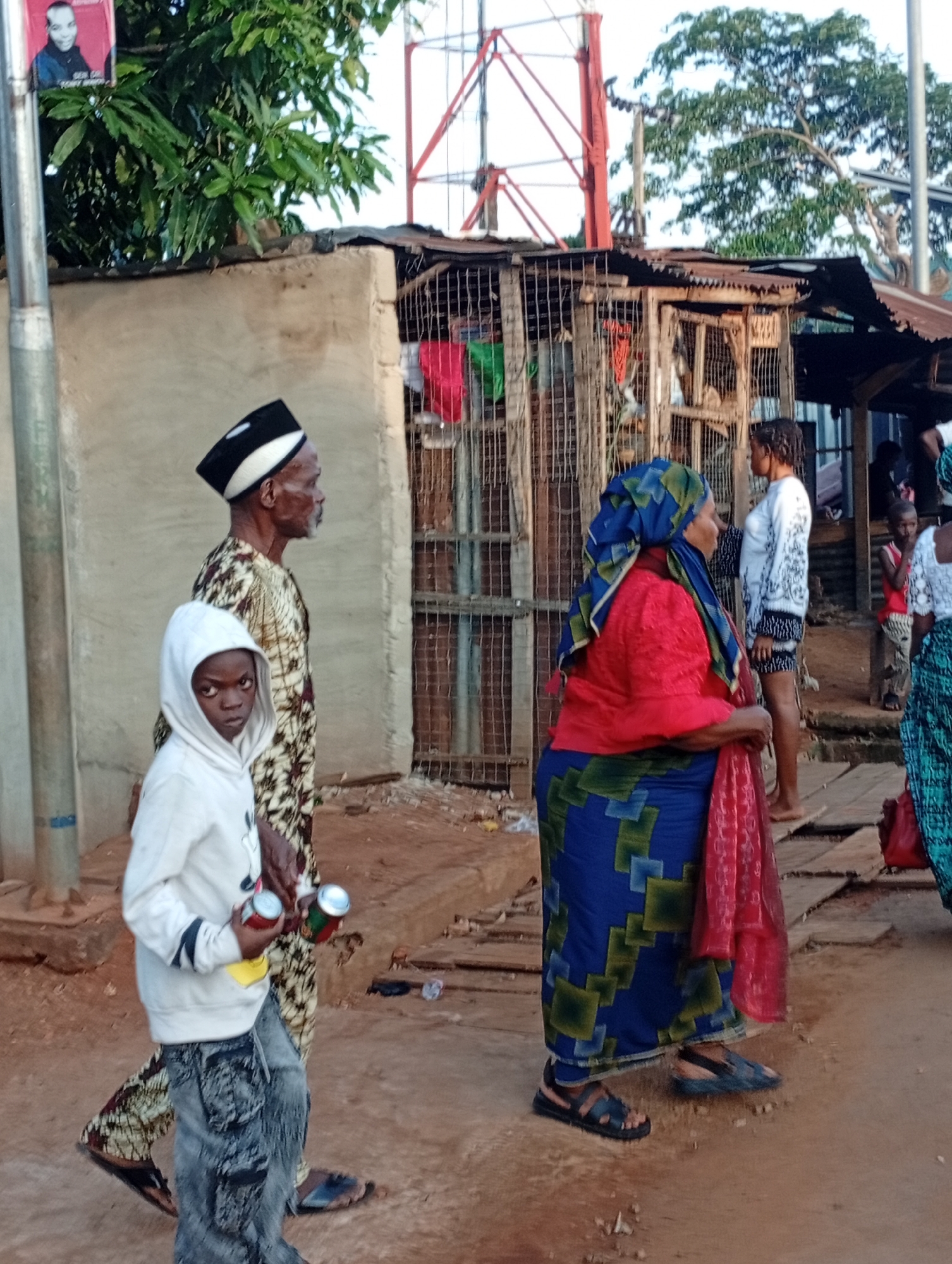 In Nsugpe, Nigeria, a street scene unfolds with several individuals. A young boy, wearing a white hooded sweatshirt and distressed denim pants, walks in the foreground, holding two cans. Beside him, an elderly man, dressed in a patterned traditional outfit and a black and white cap, walks in profile. Further back, a woman in a red top and a blue and green patterned skirt, with a red headscarf, stands looking to the side. Behind her, another woman in a white top with black detailing and dark shorts is visible. A young child in a red and white top stands in the background. The environment includes a simple structure with a corrugated iron roof and barred openings, a concrete wall, and a tall red and white telecommunications tower in the distance. Trees with green foliage are present. The ground is dusty earth and some wooden planks.