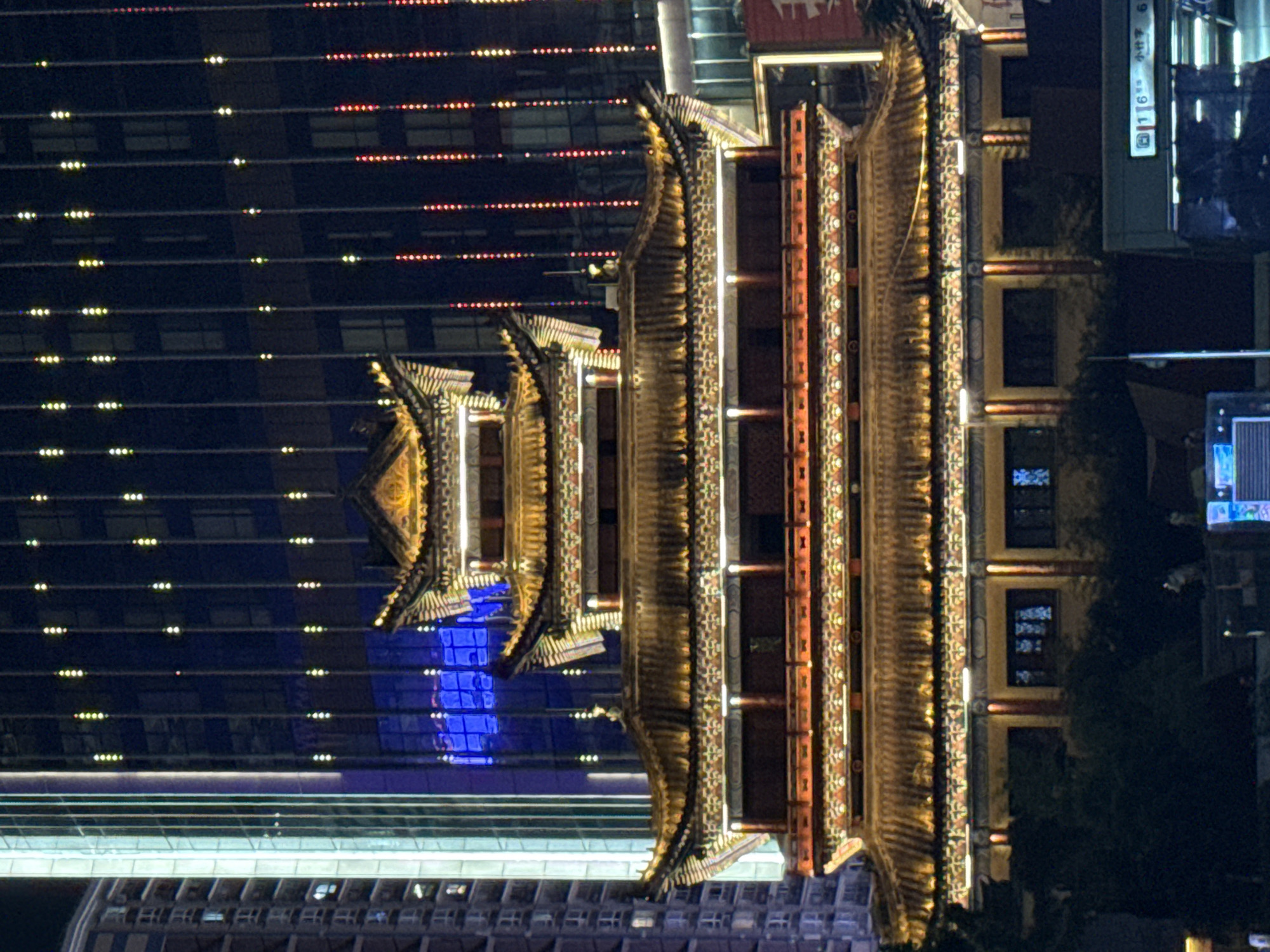 The photograph captures a brightly lit, multi-tiered traditional Chinese building at night, set against the backdrop of modern skyscrapers in Yuzhong District, China. The traditional building, adorned with intricate golden eaves and colorful decorative patterns, features several stories with illuminated windows. Its architecture is distinctly East Asian, with sloping roofs and ornate detailing.
Behind and to the left of the traditional structure, contemporary high-rise buildings dominate the scene. These skyscrapers are characterized by their glass facades, reflecting the ambient light and displaying patterns of vertical lights. One skyscraper to the left is illuminated with a cool blue light, creating a stark contrast with the warm, golden glow of the traditional building. The background is further illuminated by strings of vertical lights, some white and yellow, and others red, arranged in vertical columns against the dark night sky.
In the foreground, the lower portion of the image is shrouded in darkness, with hints of dark green foliage obscuring the base of the traditional building. At the very bottom, the corner of a green public transportation station sign is visible, bearing Chinese characters and numbers, suggesting its location within an urban transit hub. The overall impression is a juxtaposition of historical architecture and modern urban development, illuminated by a vibrant display of artificial lights.