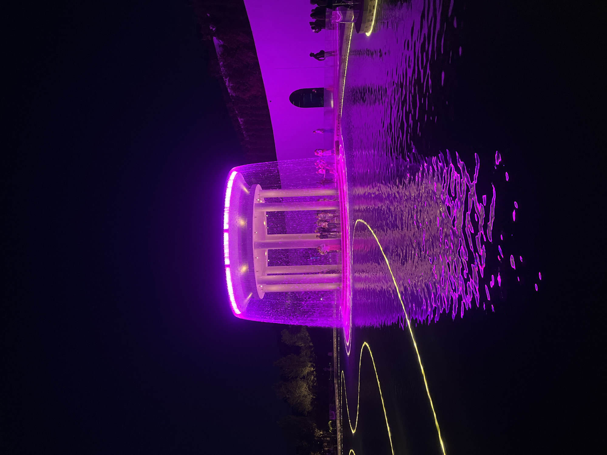 The image depicts a nighttime scene at an outdoor attraction, likely a park or waterfront area, in Krasnodar, Russia. The primary subject is a modern fountain illuminated with vibrant purple and yellow lights, which are reflected on the dark water. The fountain itself appears to be a multi-tiered structure with a cylindrical, open-air design, featuring vertical supports and a circular rim emitting the purple light. Water cascades down from the upper levels, creating a shimmering effect.
Several people are visible in the image. Some are standing on a walkway or platform surrounding the fountain, observing the spectacle. Others appear to be inside or near the base of the fountain structure. The figures are silhouetted against the bright lights, and their presence adds a sense of scale and human activity to the scene.
The setting is dark, indicating it is nighttime. The water's surface is disturbed by the fountain's action and possibly a gentle breeze, creating dynamic patterns of light and shadow. In the background, a section of a building or structure is visible, also lit with purple light, and beyond that, dark trees suggest the presence of surrounding vegetation. Yellow light trails are also visible on the water's surface, creating a visually interesting pattern that complements the purple illumination.
The overall mood is one of evening entertainment and perhaps wonder, evoked by the dramatic lighting and the active water display. No visible text is present in the image.