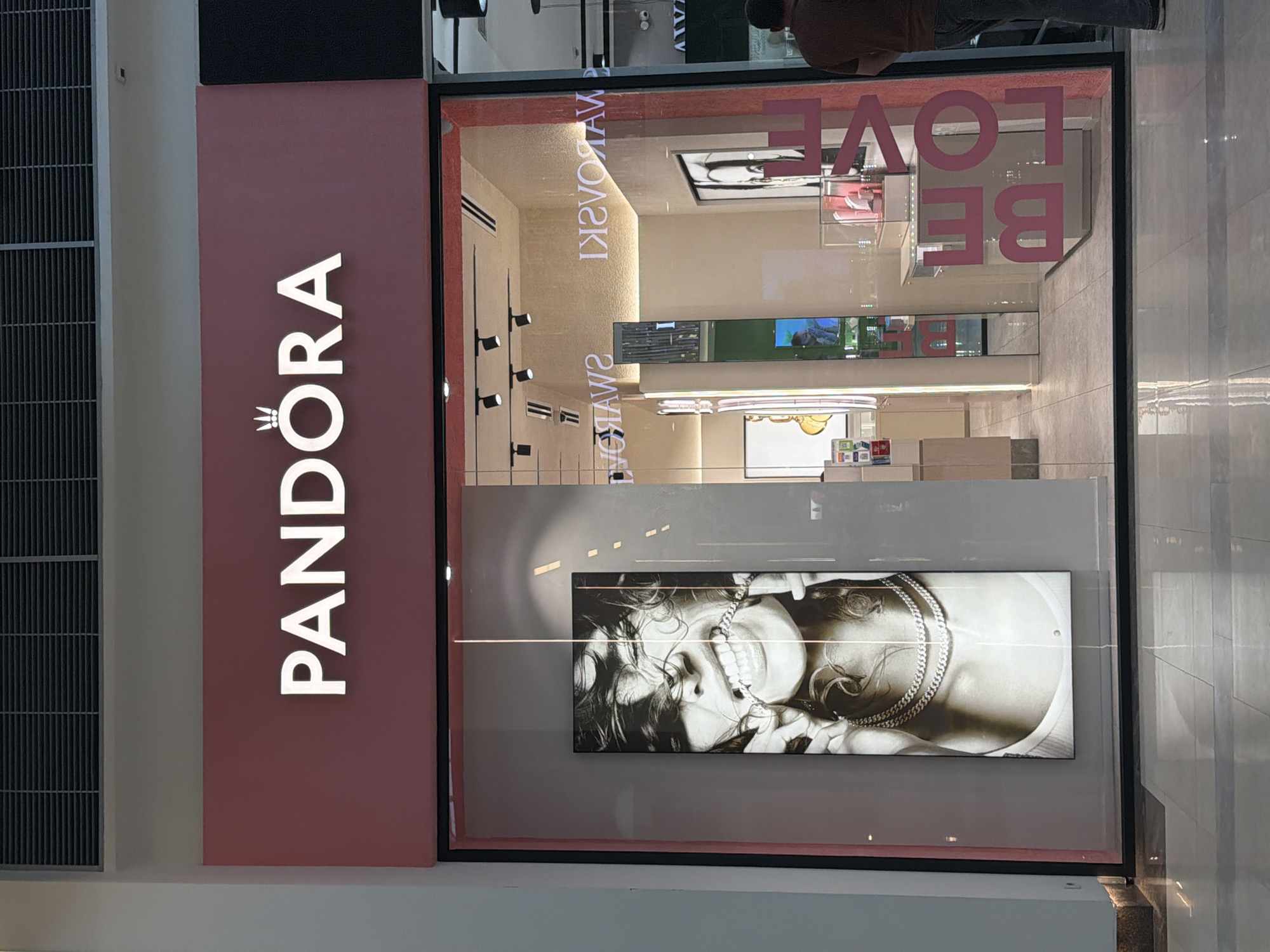The image shows the exterior of a Pandora jewelry store, likely located in a shopping mall or similar commercial space. The store's sign is prominently displayed with the brand name "PANDORA" in white, vertical lettering against a pink background. The store's window displays merchandise and an image of a woman wearing jewelry. Reflected in the glass of the store window is the image of a person, partially visible. A sign reading "BE LOVE" is also visible in the top right corner of the window. Given the signs for Swarovski, and other features of the mall, it is likely in Yerevan, Armenia.