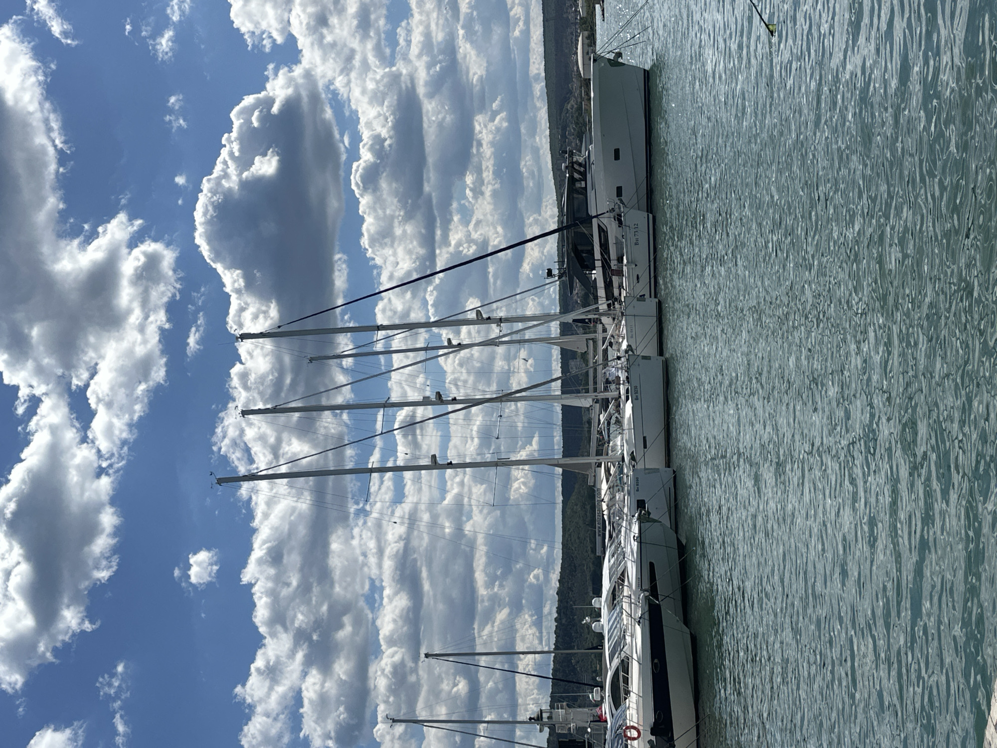 The image depicts several sailboats docked in a marina. The sky is blue with numerous white, puffy clouds. The masts of the sailboats point upwards towards the sky. The water is relatively calm with a slight ripple. In the background, there is a tree-covered landscape. The boats are white with dark windows. Although the exact location in Varna cannot be confirmed, the marina setting with sailboats and background landscape suggests a coastal or lake area within Varna.