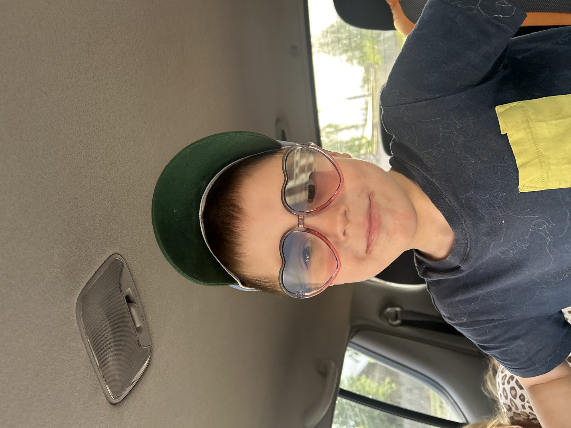 A young person wearing a dark t-shirt with a yellow patch pocket and heart-shaped glasses is sitting inside a car. They are also wearing a green baseball cap. The car's interior is visible, including the ceiling light and window. The location is not discernible from the provided image and therefore Фоки, Russia cannot be identified.