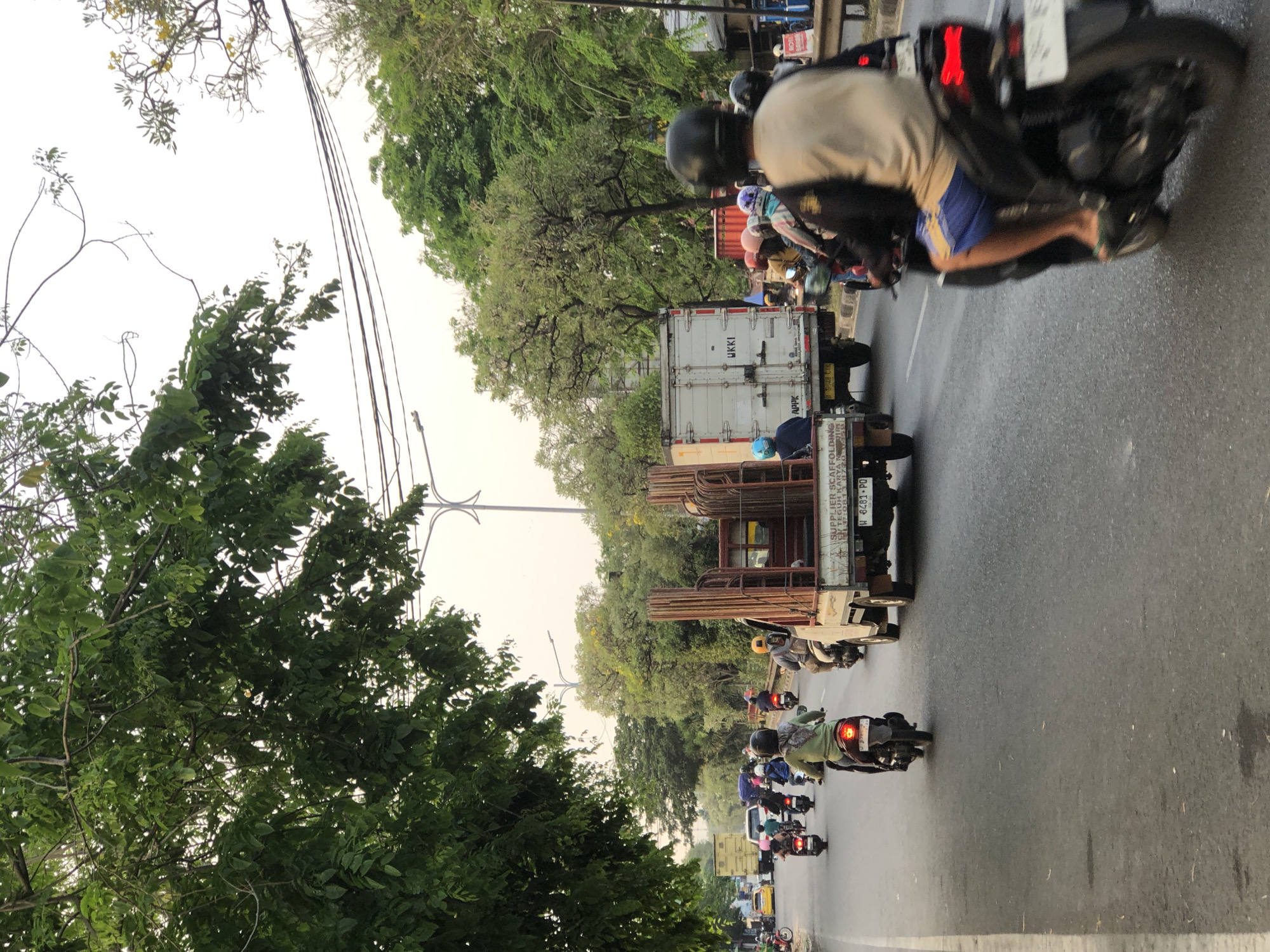 The image captures a street scene in Kludan, Indonesia, characterized by moderate traffic. A white truck carrying metal rods is prominent in the foreground, alongside multiple motorcycles and their riders. The road is paved, and trees line the sides, with some branches extending over the street. The sky appears overcast. One motorcycle is in motion, carrying a rider wearing a light-colored shirt and blue shorts, with a black helmet. Another motorcycle, to the left, carries a rider in a green top and headscarf. The truck has text on its side that includes "SUPPLIER SCAFFOLDING" and license plate "W 8481 PD". Other vehicles, including a yellow car and a larger white truck, are visible further down the road. The overall impression is of a typical day on a busy Indonesian street.