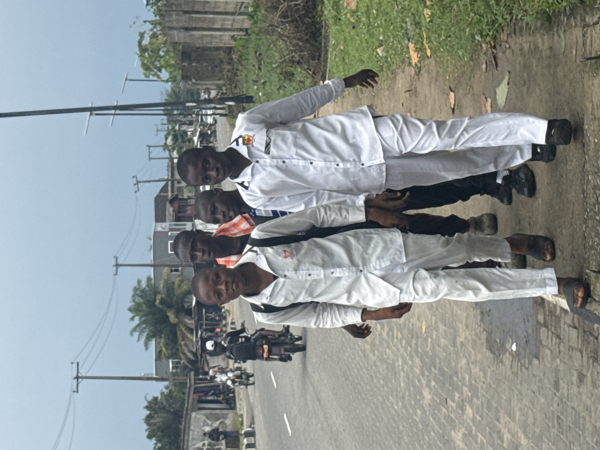 Four young men are standing on a paved road in Badagry, Nigeria. The first and fourth individuals are wearing white long-sleeved shirts and pants, while the second and third individuals are wearing casual clothing. The four men are positioned parallel to each other. Motorbikes and pedestrians can be seen in the background on the road. Utility poles and trees are present.