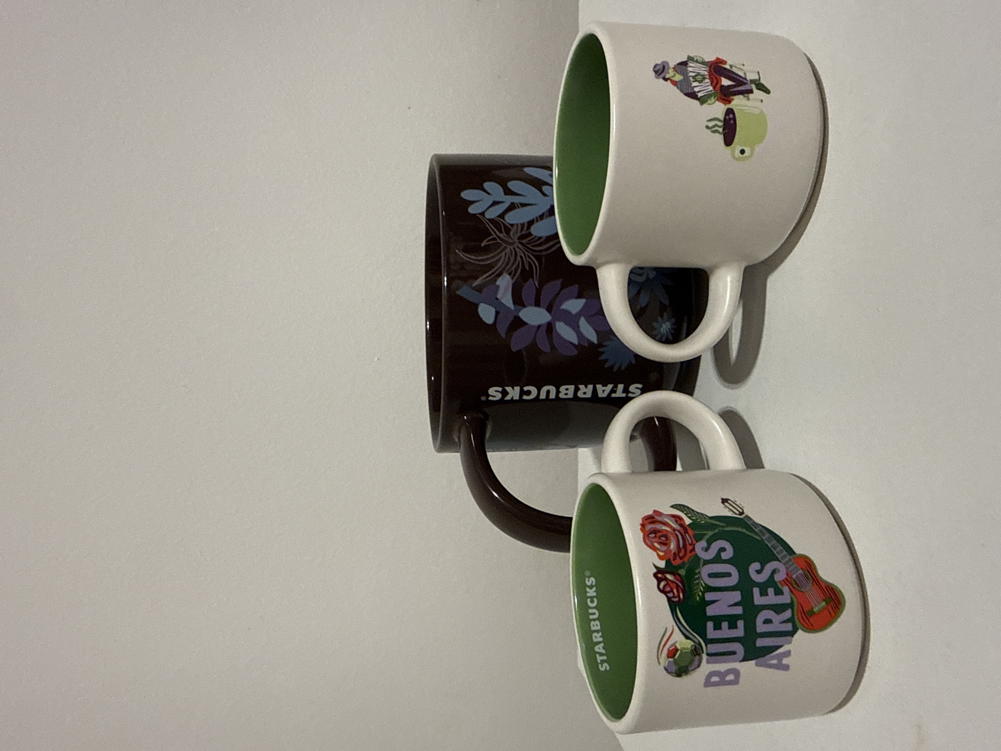 Three Starbucks mugs are displayed against a plain light-colored, possibly white, background. The image appears to be rotated 90 degrees clockwise from its original orientation.
The top mug is white with a green interior and features a colorful illustration. This design includes a character, possibly a musician, playing an accordion-like instrument, alongside a smaller green figure. The brand "STARBUCKS" is partially visible on its side.
Below and slightly behind, a dark brown mug is visible. It is adorned with a pattern of blue and purple floral or leaf motifs. The "STARBUCKS" logo is printed on its side, appearing inverted due to its placement.
The bottom mug is also white with a green interior. Its design prominently features the text "BUENOS AIRES" in purple lettering. This design is accompanied by illustrations of red roses, a green acoustic guitar, and a small soccer ball, clearly referencing the city of Buenos Aires. The "STARBUCKS" logo is visible vertically along the side of this mug.
The mugs cast soft shadows on the background, indicating a light source from the front or above. The scene depicts a static display of collectible mugs, likely part of a Starbucks city mug collection.