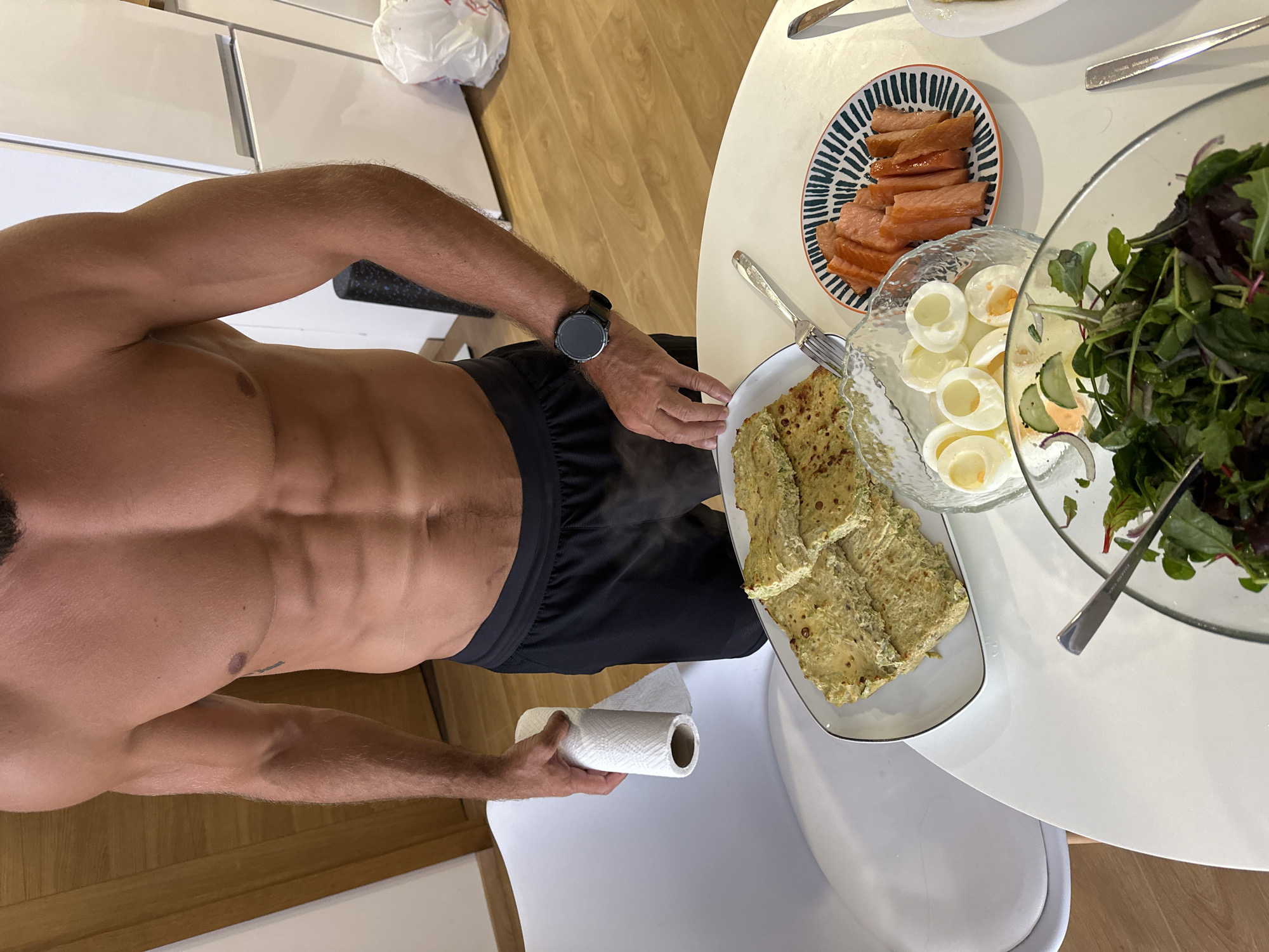 A shirtless, muscular man stands in a kitchen, holding a roll of paper towels. He is wearing black athletic shorts. On the white table in front of him are several plates of food: sliced savory pastries, a bowl of sliced hard-boiled eggs, a small plate of smoked salmon slices, and a salad in a clear bowl. In the background, a refrigerator is visible. The floor is light-colored wood. The environment is likely a domestic kitchen. The image does not contain any specific information that would suggest it is located in Donji Pobori, Montenegro.