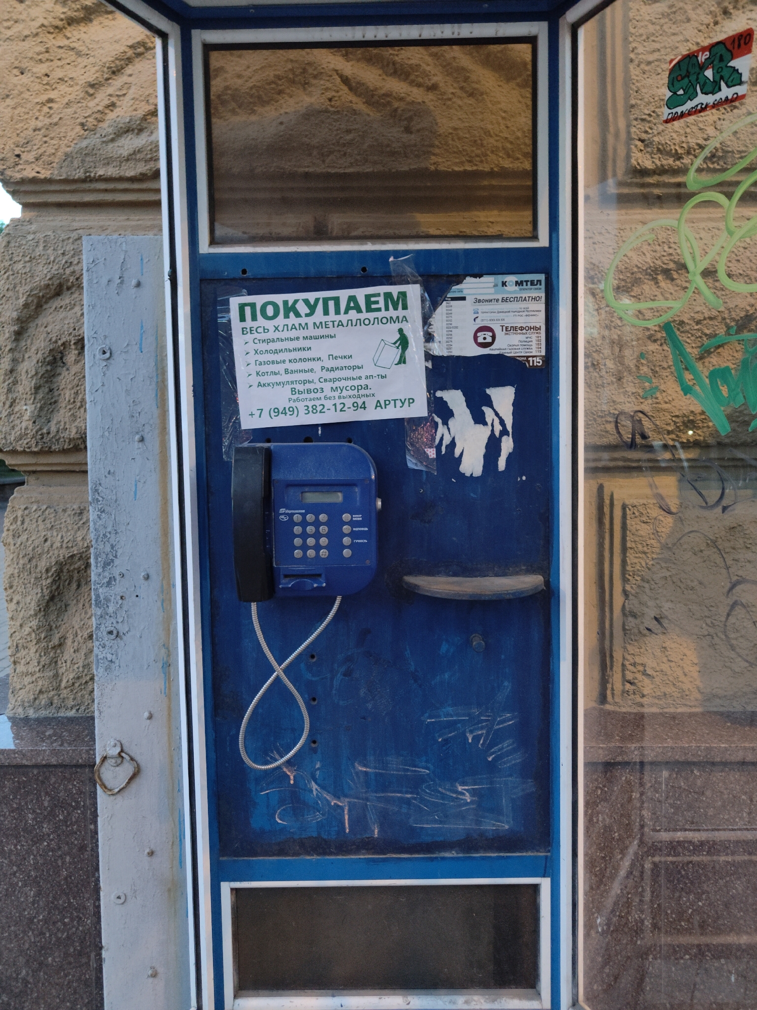 A weathered blue public telephone booth stands against a textured stone building in Donetsk, Ukraine. A faded poster inside the booth advertises scrap metal collection for washing machines, refrigerators, stoves, radiators, and batteries, with a phone number provided for services and a note indicating it operates without days off. A blue rotary phone with a coiled cord is mounted on the booth's interior. Graffiti in green and red spray paint is visible on the exterior of the booth and the adjacent building. The overall scene suggests an urban environment that has experienced wear and tear.