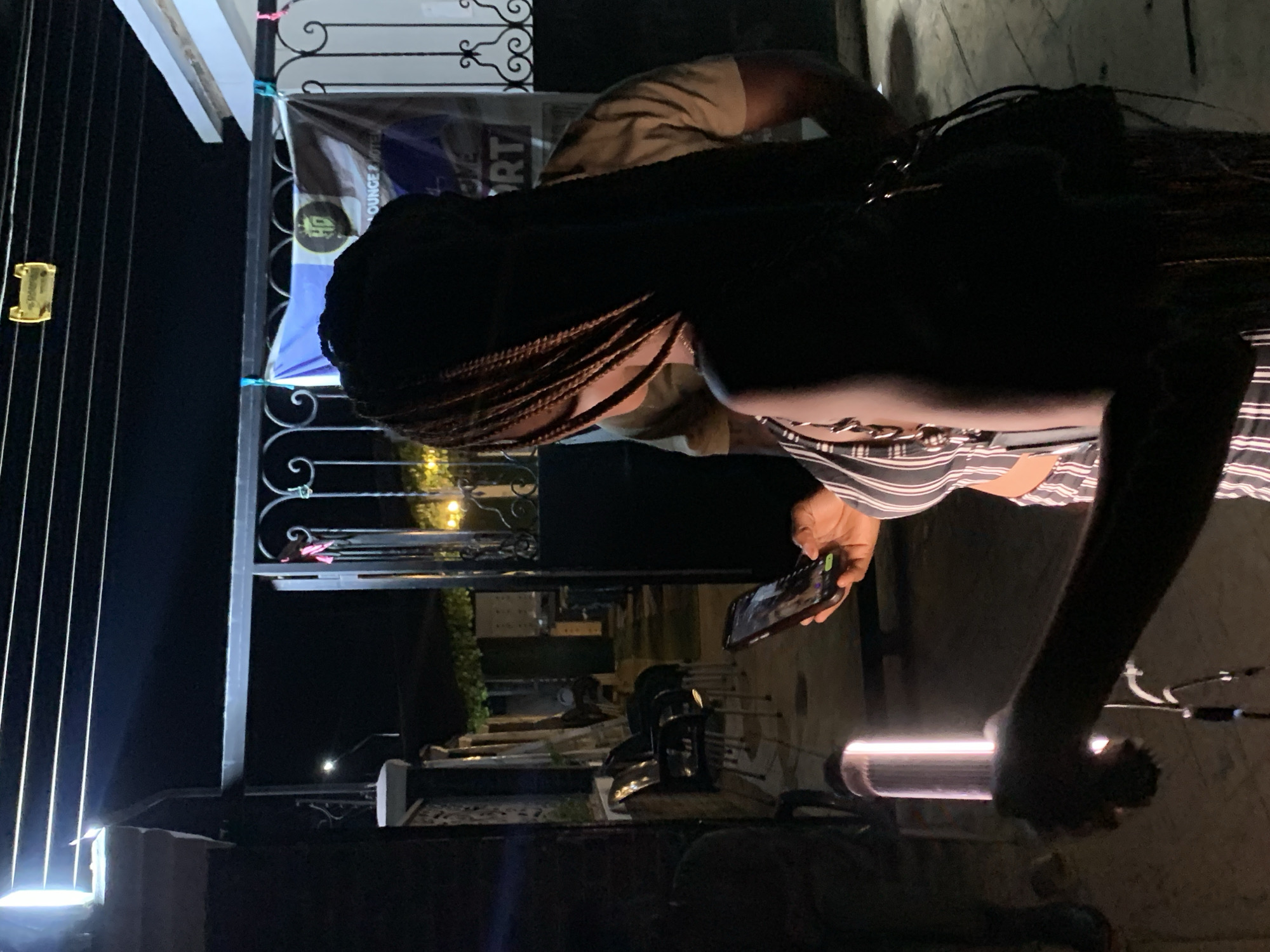 This media file depicts an evening or nighttime scene, likely outdoors or in a semi-enclosed space in Ikorodu, Nigeria. The primary subject is a person, possibly a woman with braided hair, seen from behind and to the side, focusing intently on a brightly lit smartphone held in both hands. She is wearing a striped top. Partially visible behind her is another individual's arm and shoulder, clad in a light brown shirt.
The setting features prominent artificial lighting, including a light source on the left emitting strong vertical beams and a glowing cylindrical light fixture on the right. In the mid-ground, a decorative black metal gate with ornate scrollwork is visible. A banner hangs from this gate, partially displaying text that reads "OUNGE & HOTEL" and "RT," suggesting it pertains to a "Lounge & Hotel" or a "Sport" related establishment. Further in the background, faint outlines of tables and chairs can be discerned, indicating a seating area typical of a lounge, bar, or event venue. The overall atmosphere is dark, illuminated by various artificial light sources.
