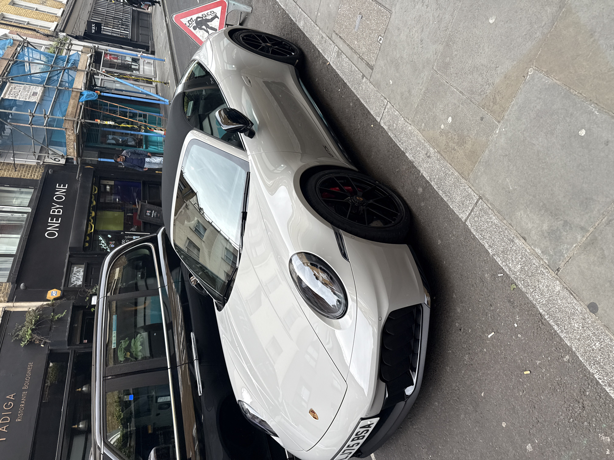 The image depicts a daytime street scene in an urban area, likely London given the visible cues. A light-colored Porsche sports car, possibly white or light grey, is parked prominently on the side of a paved road. Its license plate reads "LJ75 BSA". The car features black multi-spoke wheels with visible red brake calipers. Behind the Porsche, a black car is also parked.
To the left, multi-story buildings line the street. One building features extensive scaffolding with blue netting, suggesting ongoing construction or renovation. Text on the netting includes "ABILITY HEAVY-DUTY EWCS LTD" and a phone number. Several shop signs are visible: "ONE BY ONE", "ADIGA RISTORANTE BOLOGNESE", and "WAX Tattoo". A person in a dark jacket is standing near the scaffolding.
On the paved sidewalk, a red triangular warning sign with two stylized black figures (often indicating a school crossing or similar pedestrian hazard) is visible. The street surface is asphalt, and the sidewalk consists of large grey paving slabs. Scattered cigarette butts are visible on the street near the front of the Porsche. The lighting suggests an overcast sky, with no harsh shadows.