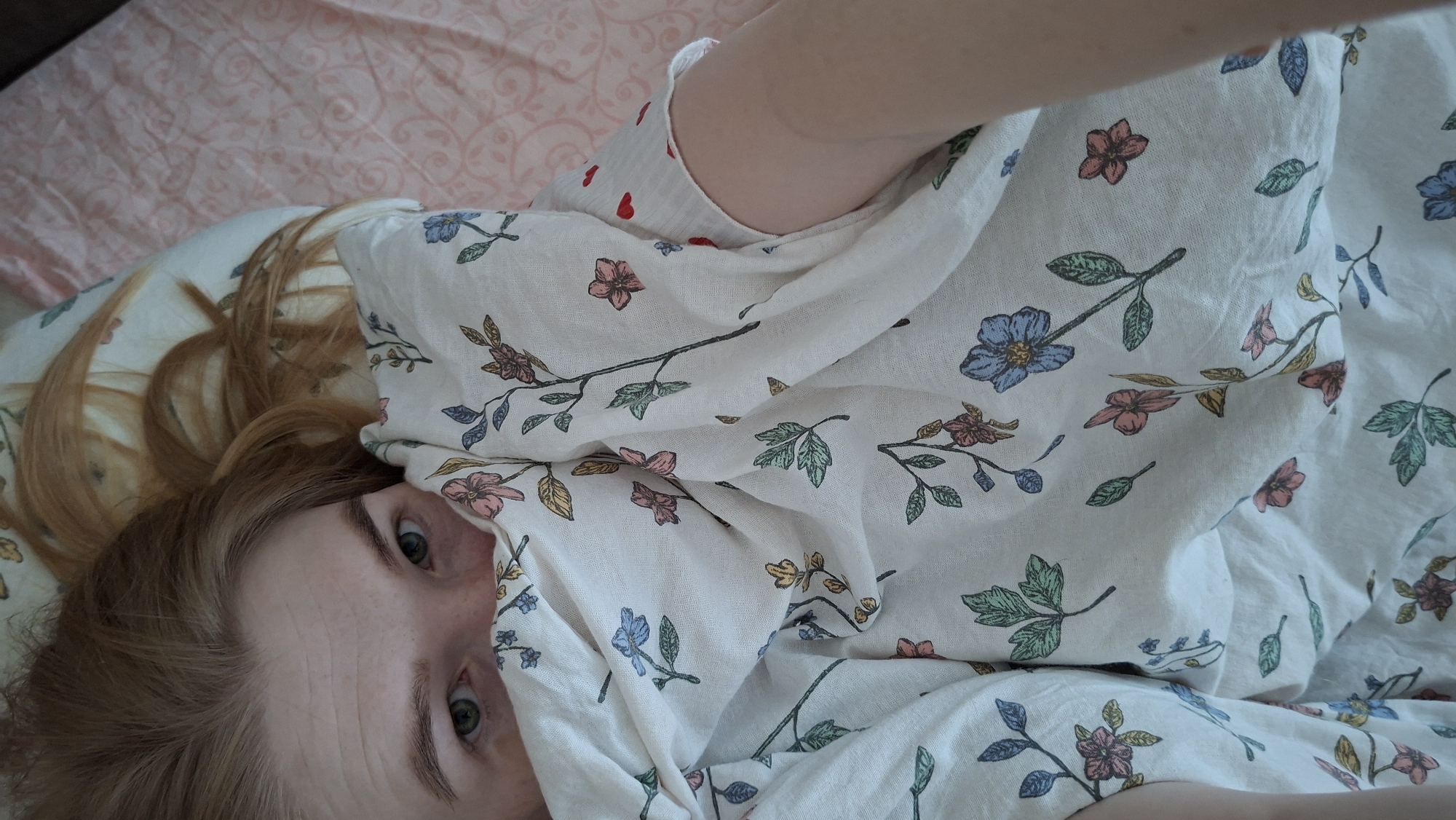 A woman with reddish-blonde hair lies on a bed, partially covered by a floral-patterned sheet. Her eyes are visible. The background shows a pillow and patterned bedding. The location is Chaikovsky, Russia.