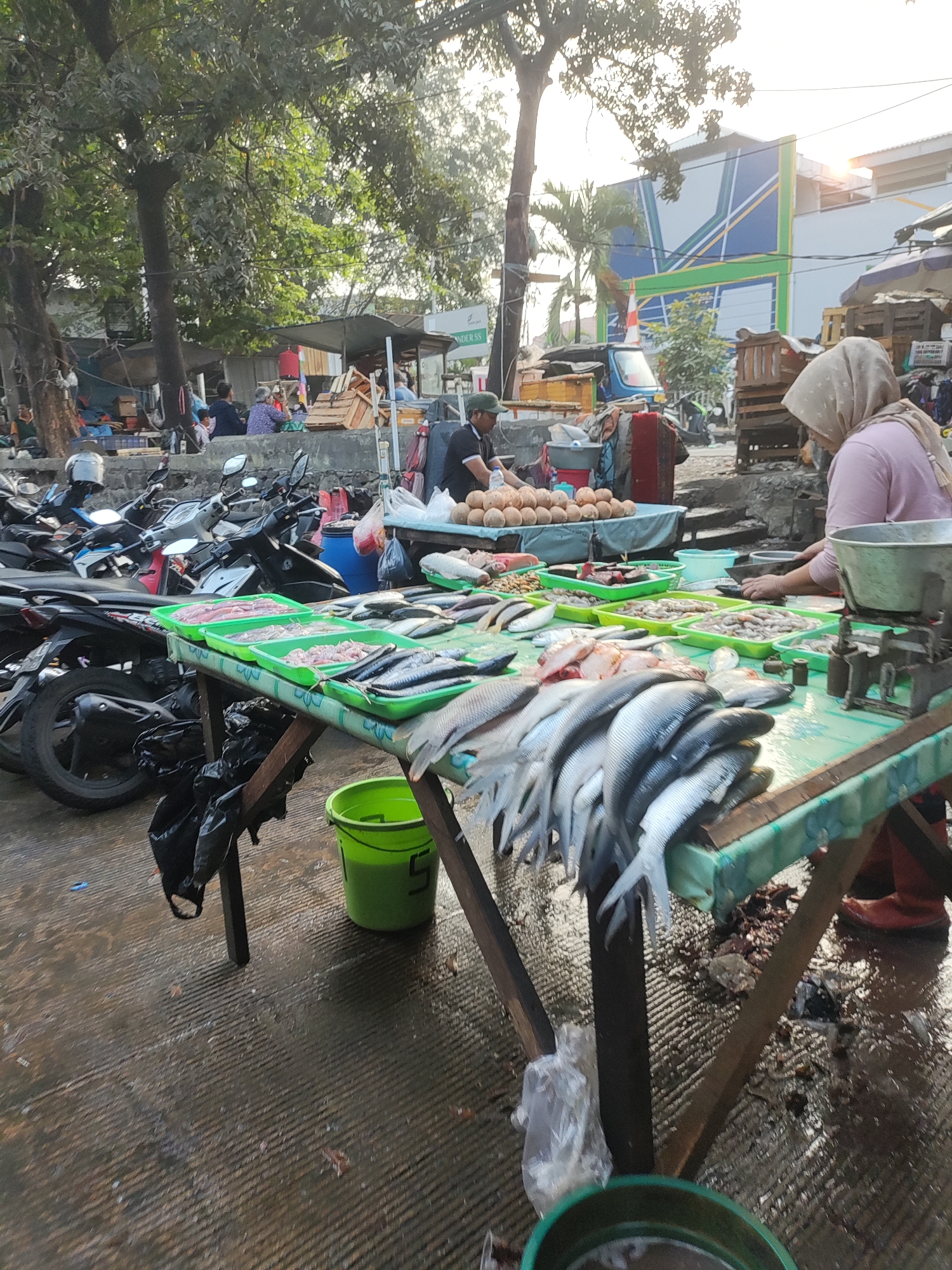 This image depicts a busy fish market scene in East Jakarta, Indonesia, likely on a street or open-air market area given the presence of parked motorcycles and the general setting. In the foreground, a table laden with various types of fresh fish is displayed, with trays of fish, some whole and some filleted, arranged for sale. A woman wearing a hijab is visible on the right, tending to the fish. Behind the fish stand, more people are seen, some appearing to be customers or vendors, in the background. The environment appears somewhat damp, with water or wet surfaces on the ground. The overall atmosphere is that of a lively marketplace with goods and activity.