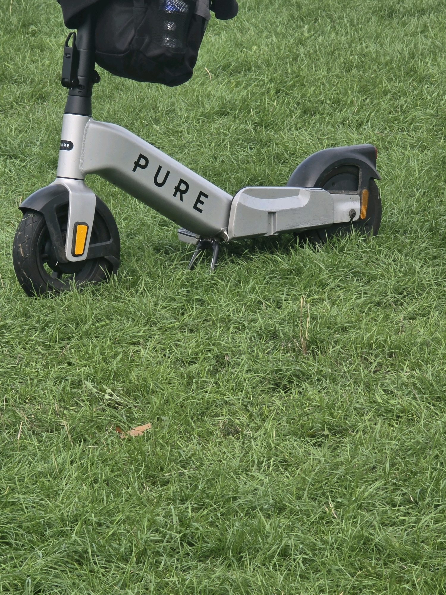 An electric scooter is parked on a grassy area. The scooter is silver and has the brand name "PURE" printed in black on its frame. It has a black bag attached to the handlebars and a black fender over the front wheel. The scooter's front wheel is visible, and it has a yellow reflector. The background is a dense, green, mown lawn with some fallen leaves. The image does not provide any specific indicators of its location within East Staffordshire, United Kingdom.