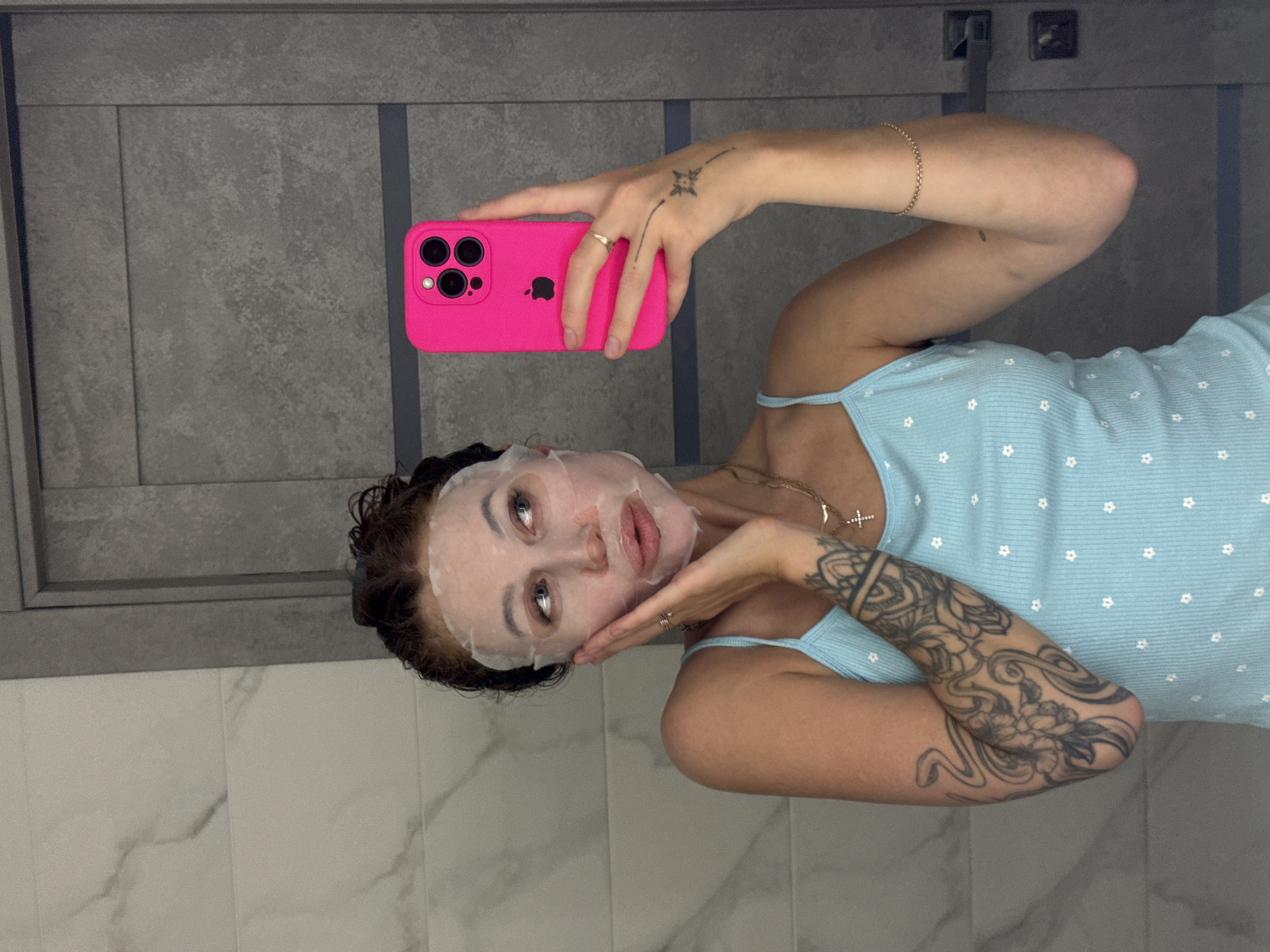 A young woman is captured in a mirror selfie, showing her engaged in a skincare routine. Her face is covered with a white sheet mask, through which her eyes and lips are visible. She has dark hair, which appears to be damp or styled away from her face, and she is making a slight pouting expression. She is wearing a light blue ribbed camisole or tank top adorned with small white floral patterns.
She holds a bright pink iPhone in her right hand, which is equipped with a distinct triple-lens camera module, suggesting an iPhone 14 Pro or Pro Max model. A gold ring is visible on her right hand's ring finger, and a small, stylized star tattoo with a dotted line extends from the back of her hand towards her wrist. Her right wrist also features a thin gold chain bracelet. Around her neck, she wears a delicate gold chain with a small cross pendant. Her left arm is heavily tattooed with intricate black ink designs, creating a full sleeve. The Apple logo is discernible on the back of the phone case.
The background of the selfie features dark grey, rectangular wall tiles with lighter grey grout lines or paneling, while the lower portion of the scene reveals lighter, possibly white or marble-patterned, tiled surfaces. This setting suggests an indoor location, likely a bathroom or a private dressing area within a residence in Chaikovsky, Russia. The overall impression is one of relaxed self-care or a playful moment captured for social sharing.