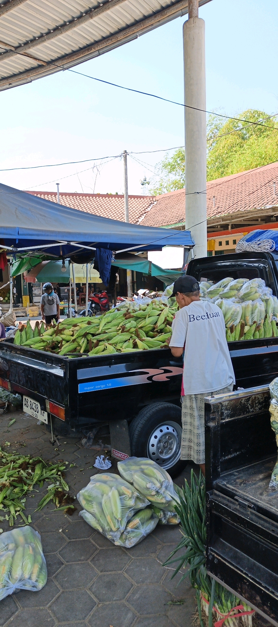 At an outdoor market in Klaten, Indonesia, a person wearing a white t-shirt with the text "BeeLuat" and patterned shorts is standing next to the open tailgate of a black pickup truck. The truck bed is overflowing with corn on the cob, some still in husks, others shucked. Several bags of corn are also visible on the ground next to the truck, as well as discarded corn husks. In the background, a blue tarp-covered stall is set up, with various items hanging from it. Other people and a red motorcycle are partially visible under the stall. The market appears to be covered by corrugated metal roofing supported by poles.