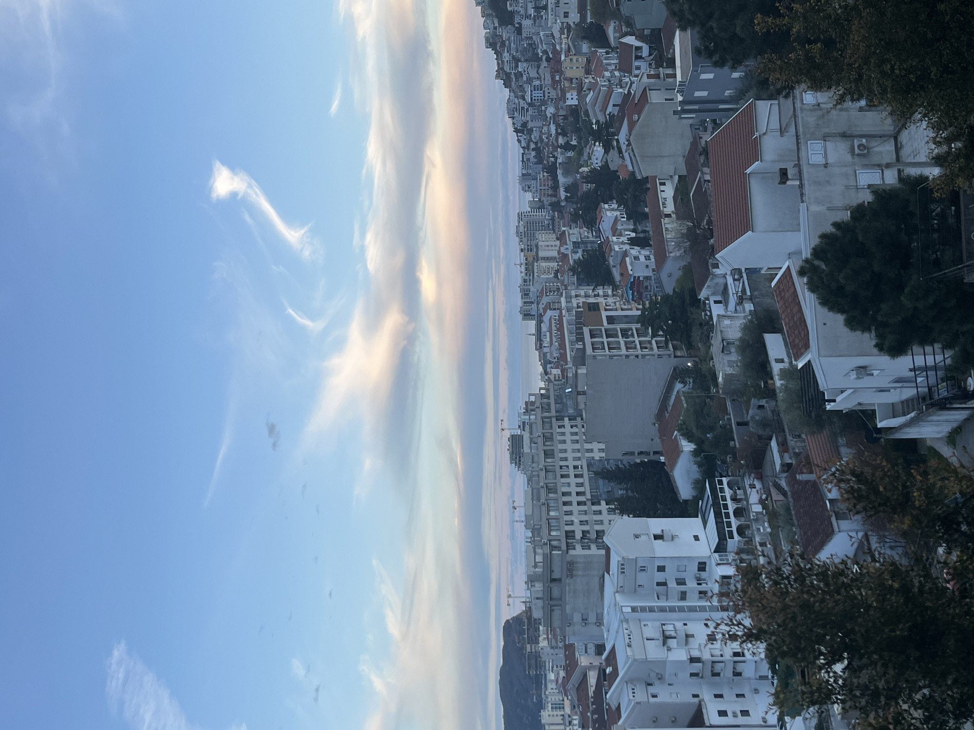 The image shows a vertical cityscape in Budva, Montenegro, viewed from a high vantage point. The sky transitions from a clear blue to light pastel colors with wispy clouds. Below, a densely populated urban area with buildings of varying heights, predominantly white or gray with some red-tiled roofs, slopes towards the sea. The structures appear to be a mix of residential and commercial buildings, interspersed with green trees. In the distance, a mountain partially obscures the horizon. The buildings nearest to the camera are partially obscured by trees.