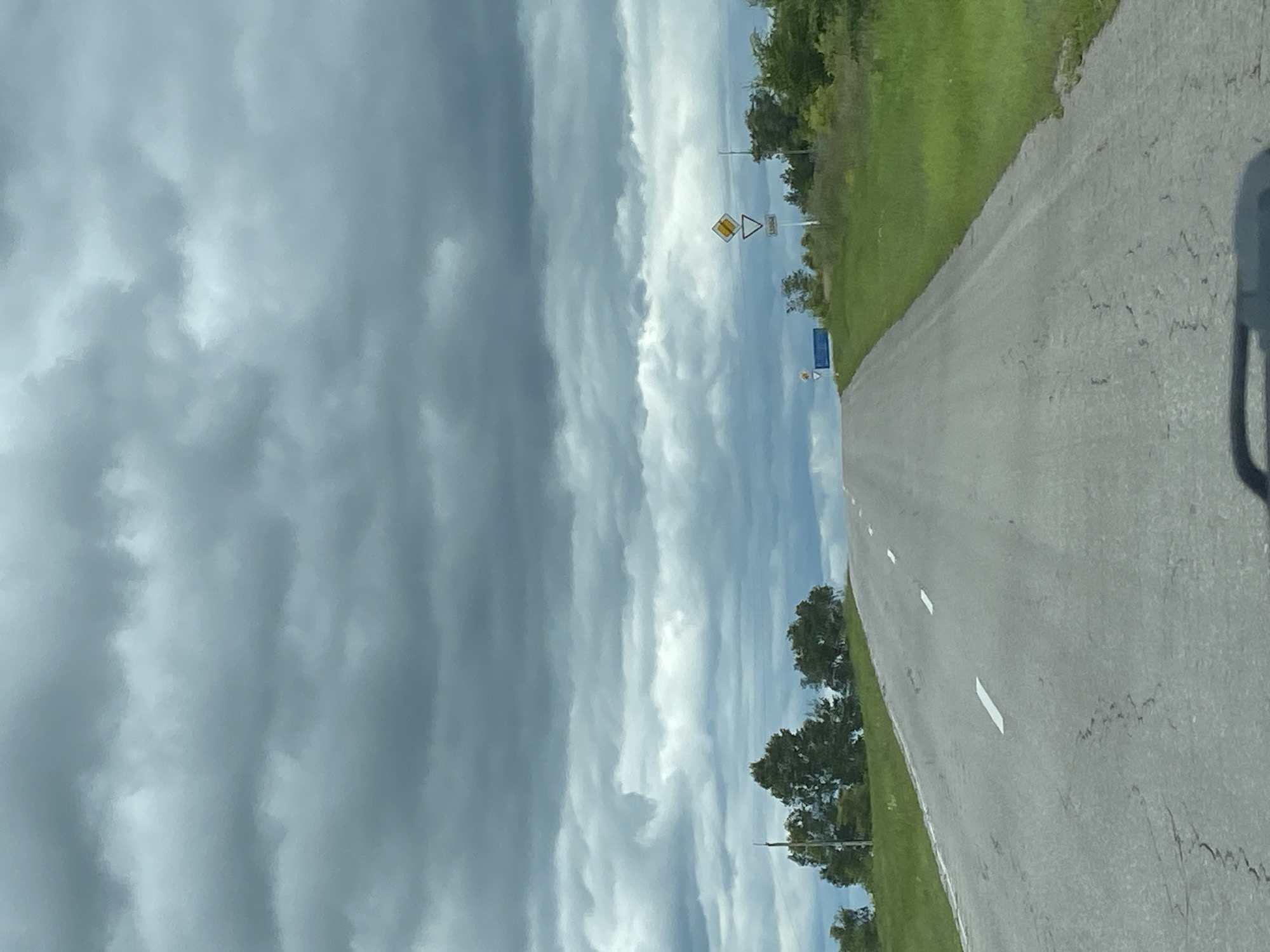 A grey, overcast sky looms over a paved road that ascends a gentle incline. White lines mark the lanes of the road, which appears somewhat worn and cracked. To the right of the road, lush green grass borders the asphalt, and a scattering of trees, predominantly green and deciduous, dot the landscape. In the distance, several road signs are visible. One sign, a yellow diamond with a black border, is accompanied by a white triangle with a black border pointing downwards, and below that, a rectangular sign with white text on a white background. Further to the left, a blue rectangular sign is positioned near a cluster of trees. The scene suggests a rural or semi-rural setting, potentially on the outskirts of a town or village. There are no people or animals visible in the image.