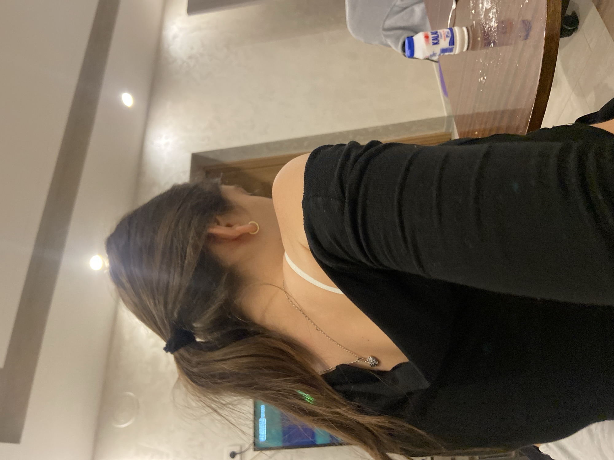 The image shows a woman with brown hair pulled back in a ponytail, wearing a black sweater and a gold hoop earring. She is also wearing a necklace. Part of a table with a bottle of "Kalak" and some white powder on it is visible in the background. There is also a TV in the background. The location is not specified, and it is unknown if it is Dragovishtitsa, Bulgaria.