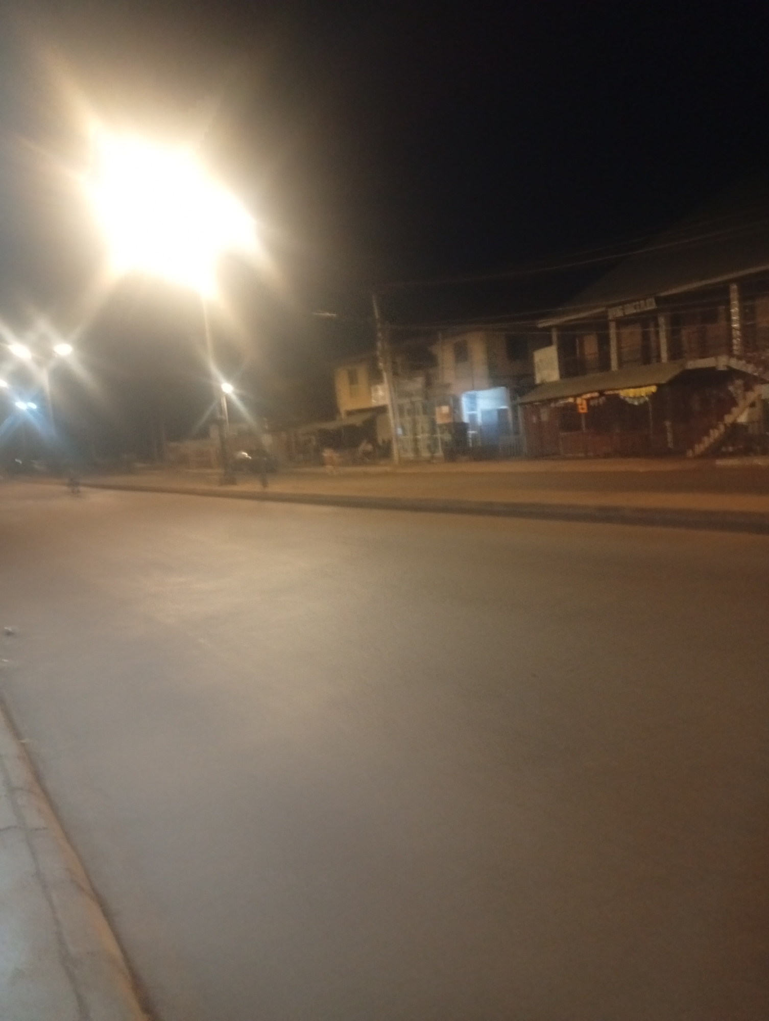 The image depicts a street scene at night in Bukuru, Nigeria, illuminated by streetlights. A bright streetlight dominates the left side of the frame, casting a strong glare. The road is paved and appears to be empty of cars, though some blurry figures and possibly motorcycles are visible further down the street. On the right side of the road, there are buildings with illuminated windows and signs, suggesting commercial establishments. The overall atmosphere is dark and somewhat deserted, typical of a nocturnal urban environment.